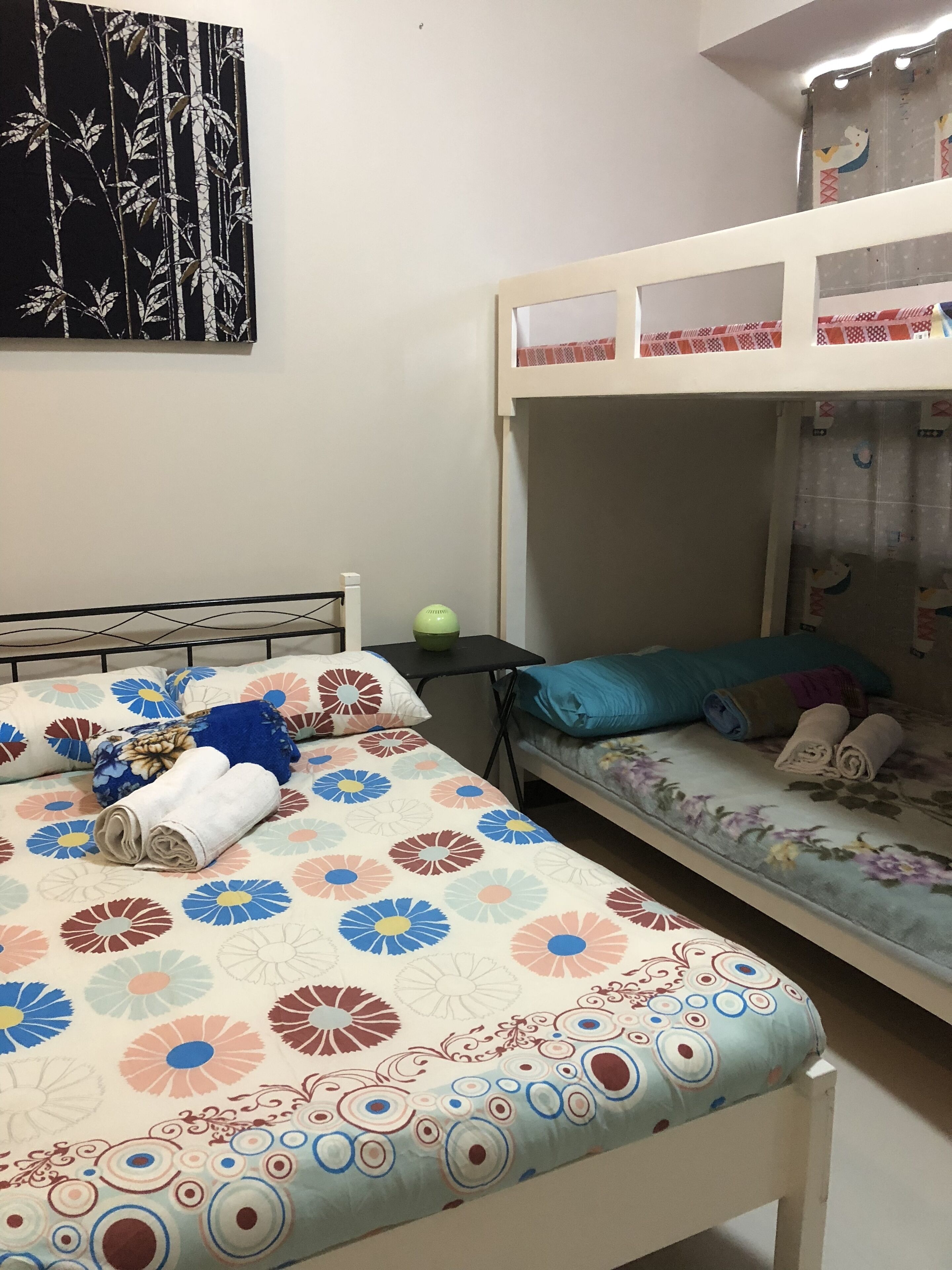 A dormitory room with two beds, one with a floral bedspread and the other with a floral pillow. There is a bunk bed above the smaller bed, and a green bowl on a small table beside the larger bed.