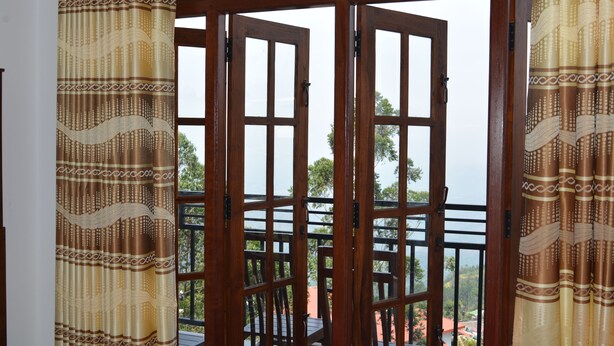 Vista a través de una ventana con marco de madera y cortinas estampadas, que muestra un balcón con barandilla y vistas a zonas verdes y edificios.