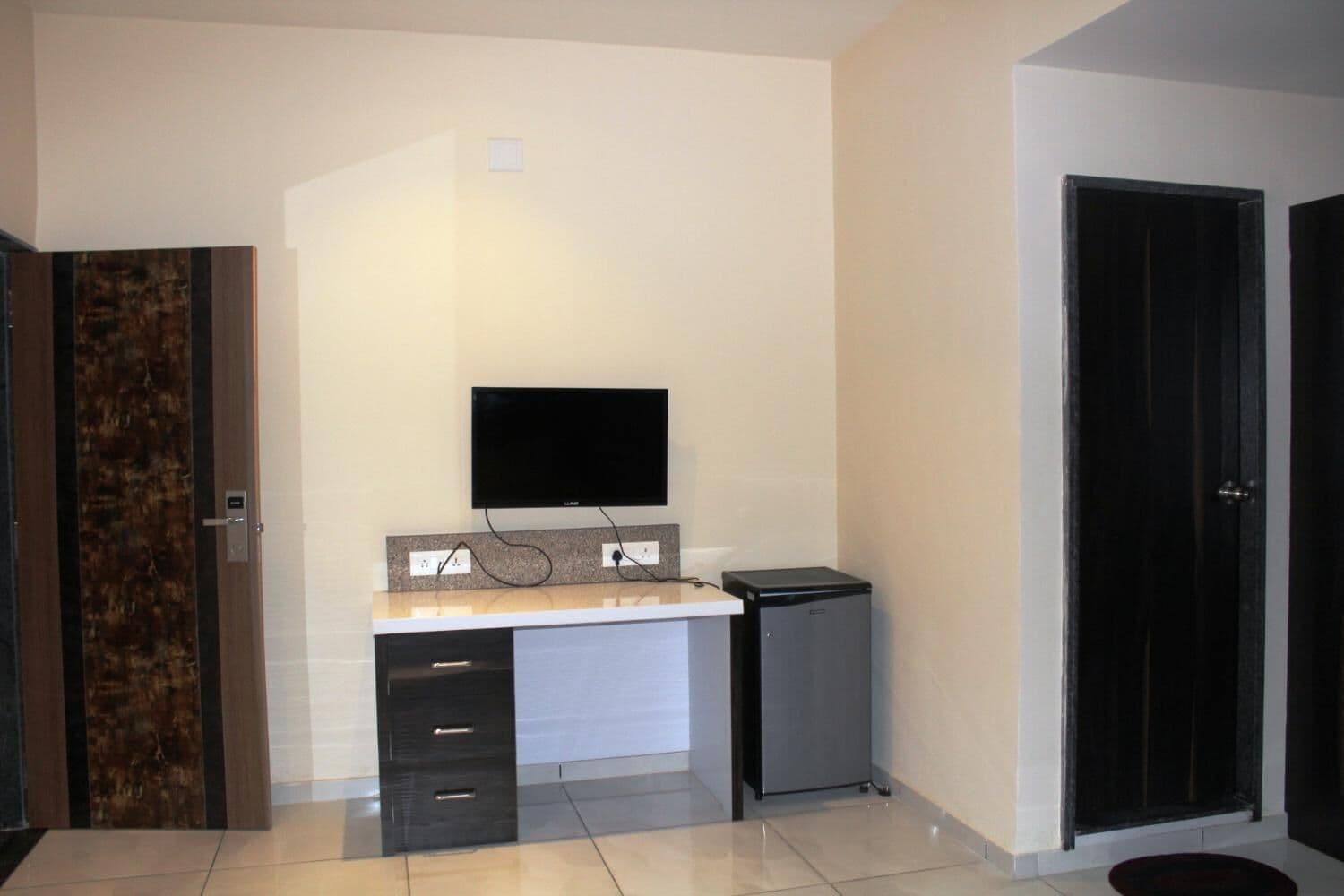 A room with a flat-screen TV mounted on the wall, a small desk with drawers, a compact refrigerator, and a door with a dark frame.