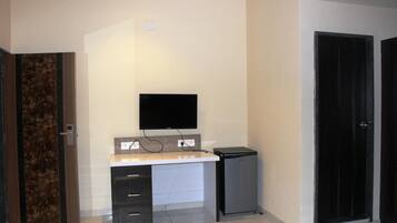 A room with a flat-screen TV mounted on the wall, a small desk with drawers, a compact refrigerator, and a door with a dark frame.