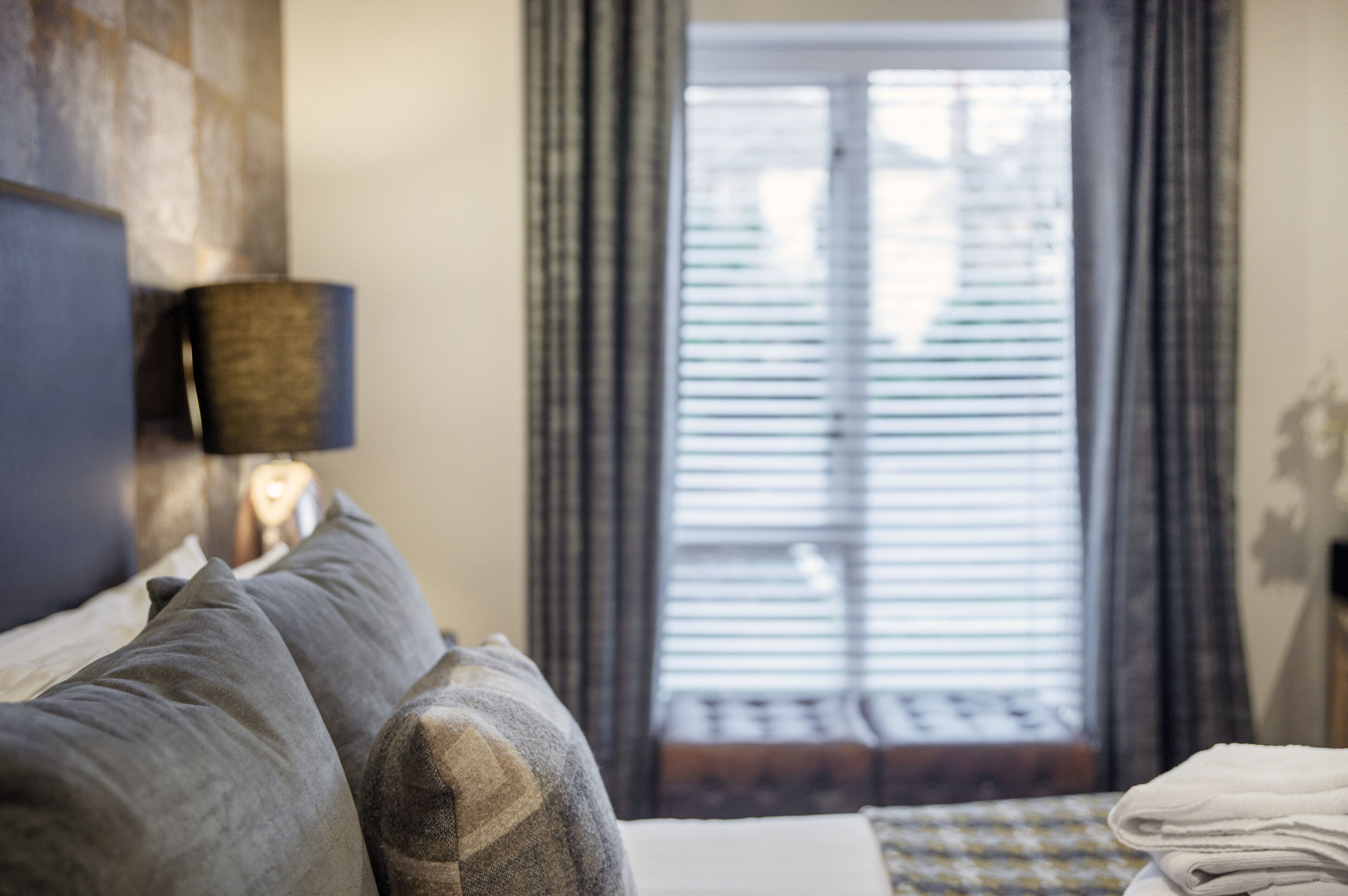 A hotel room with a bed, a lamp, a window with blinds, and a view of the outside.
