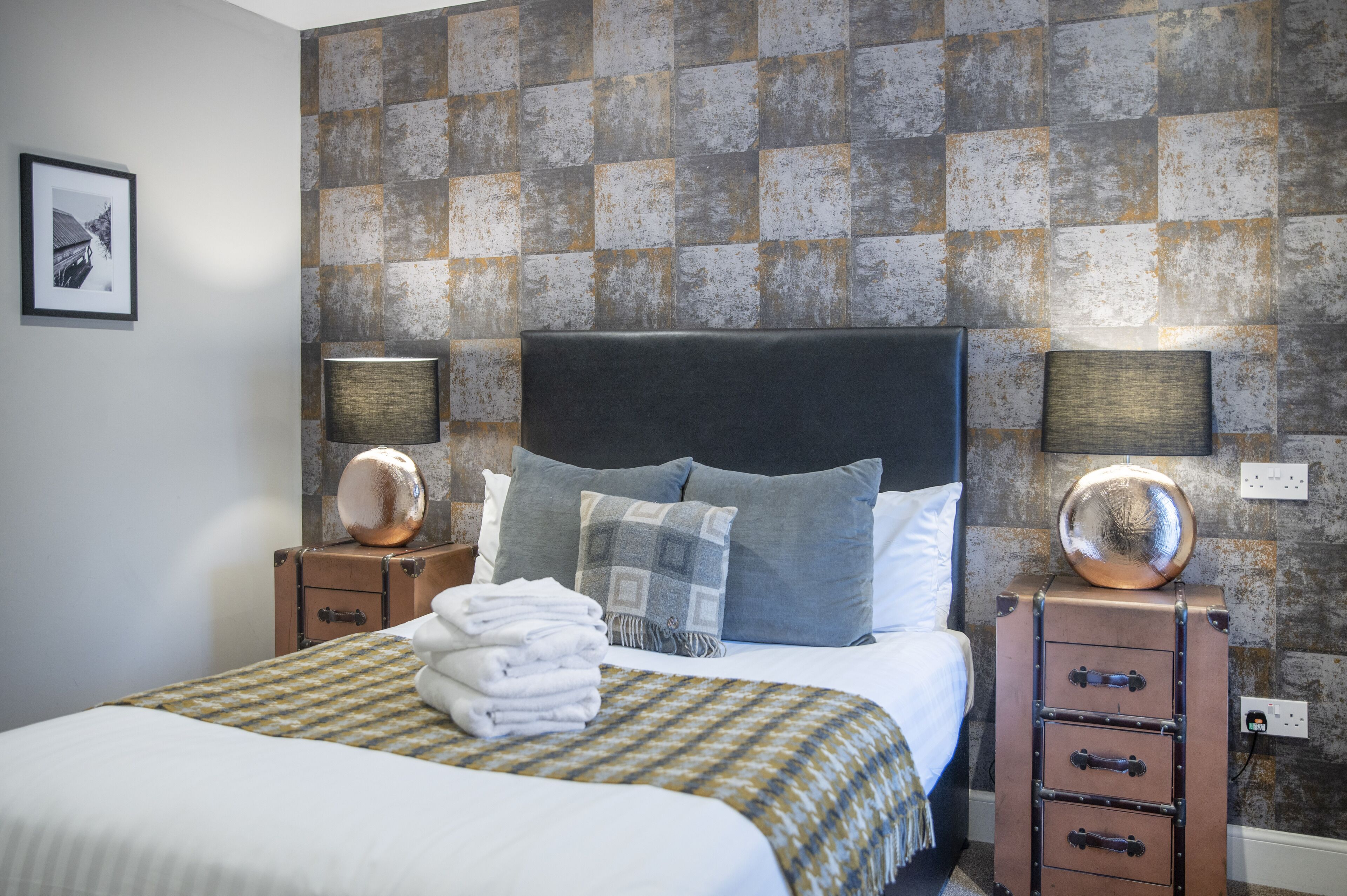 Un lit bien fait, avec une couverture à carreaux et des oreillers, une valise posée sur une table de chevet, et un mur à motifs derrière le lit.