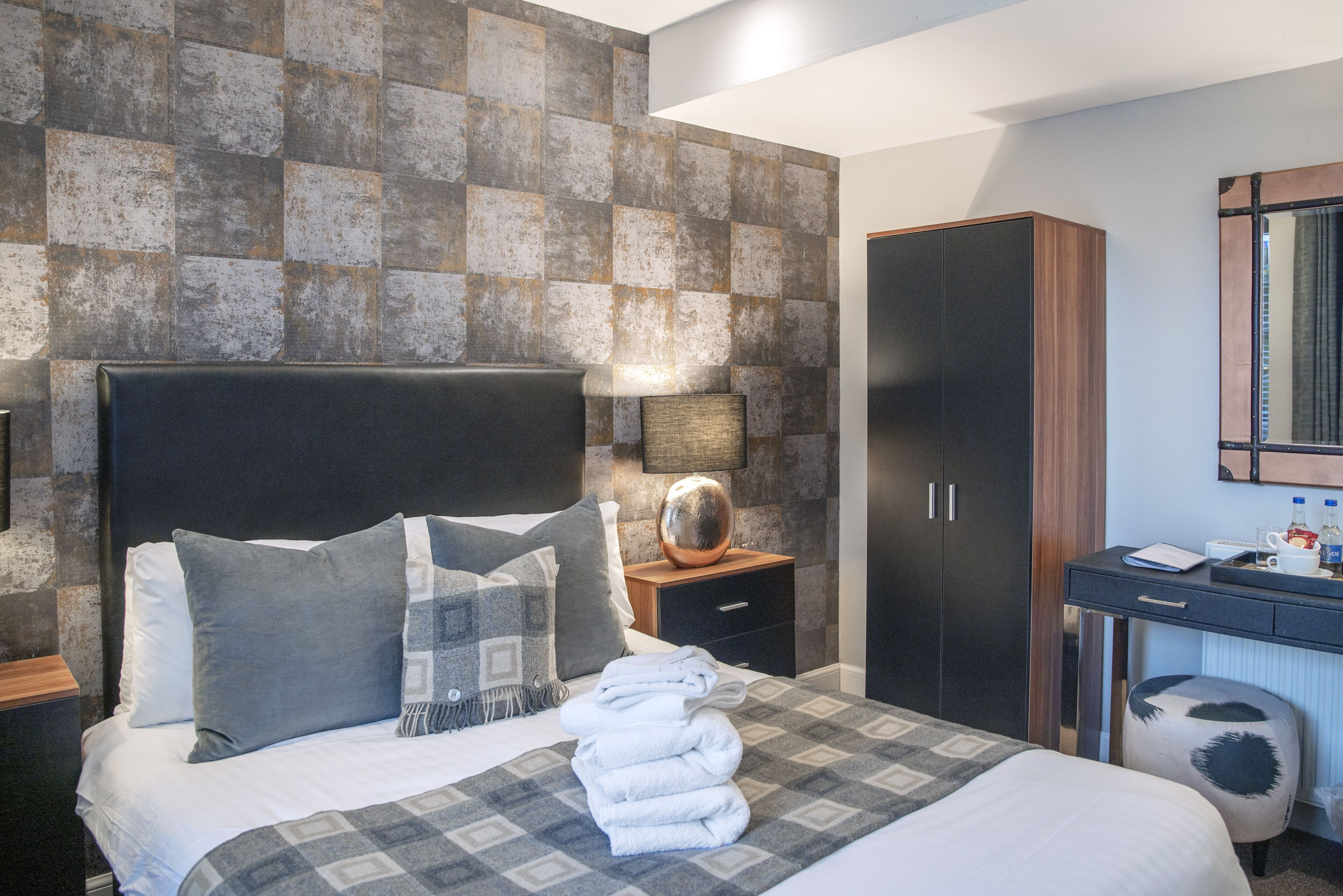 A neatly made bed with pillows and a folded towel, a bedside table with a lamp, a wardrobe, and a desk with a mirror.