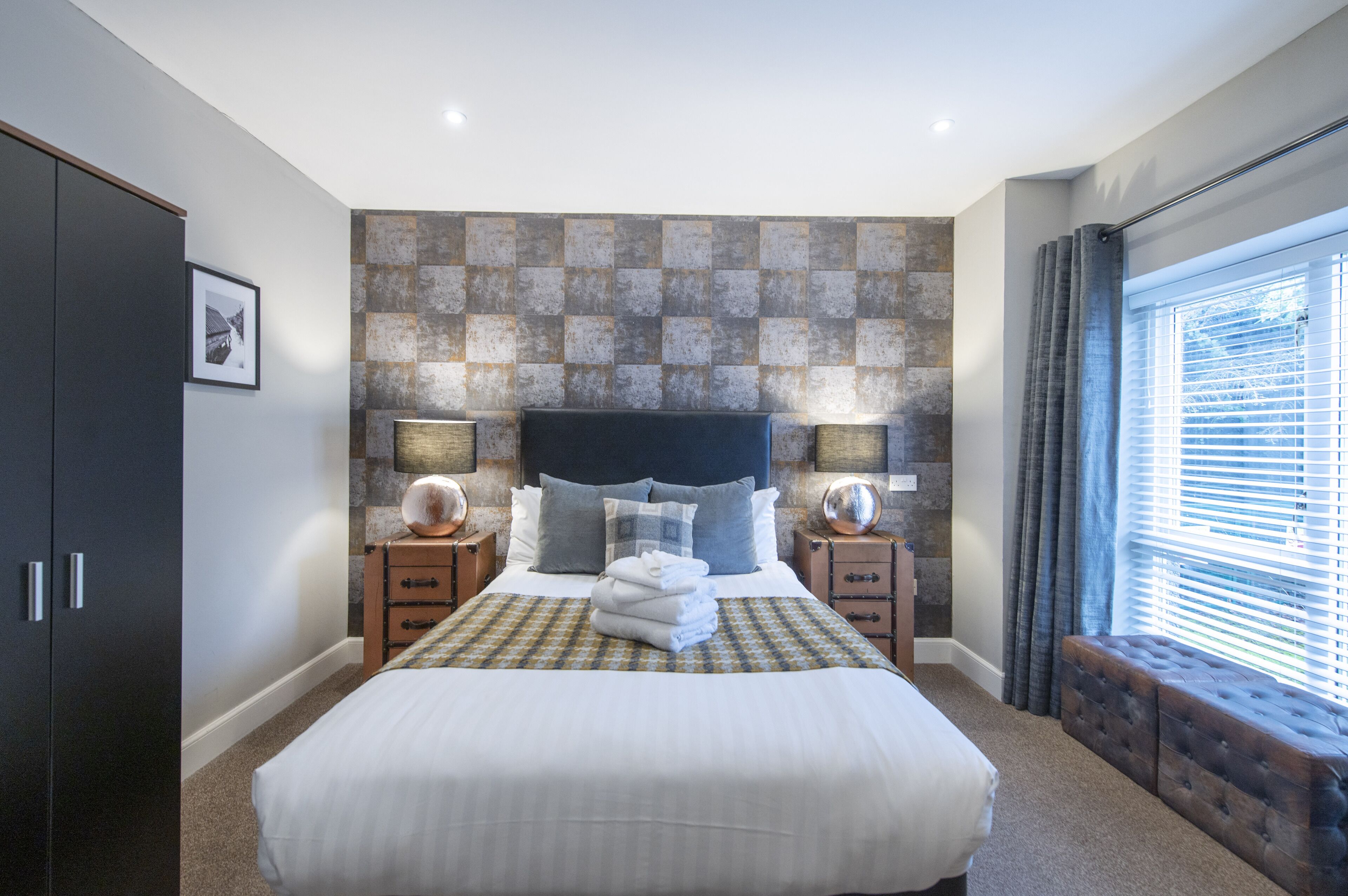 A bedroom with a large bed, two bedside tables with lamps, a headboard, a wardrobe, and a window with curtains.