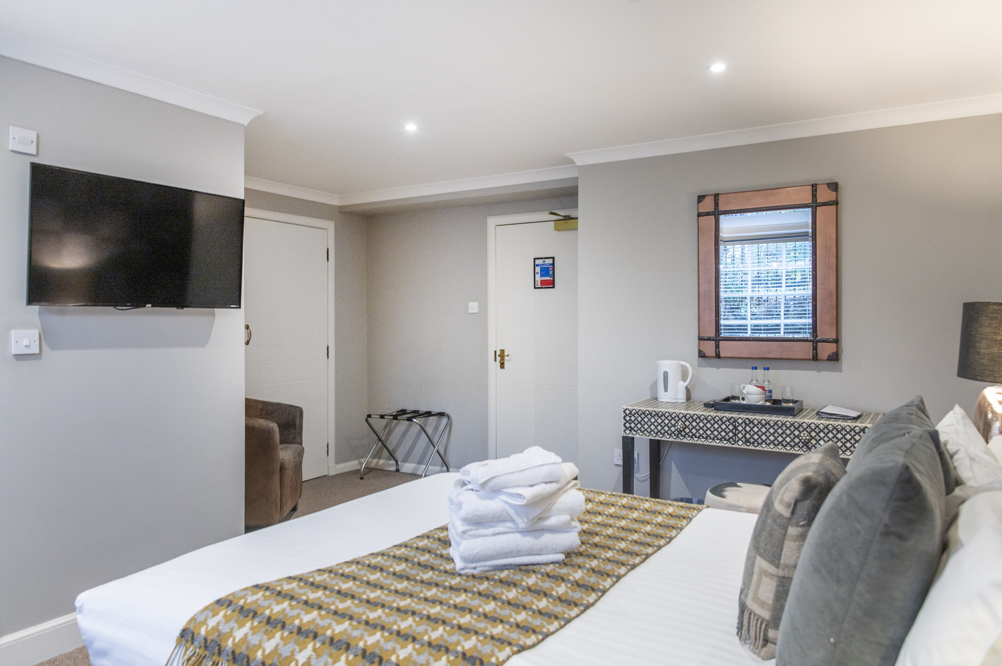 Una camera d'albergo con un letto, una TV a muro, una scrivania con una macchina del caffè e una finestra con persiane.