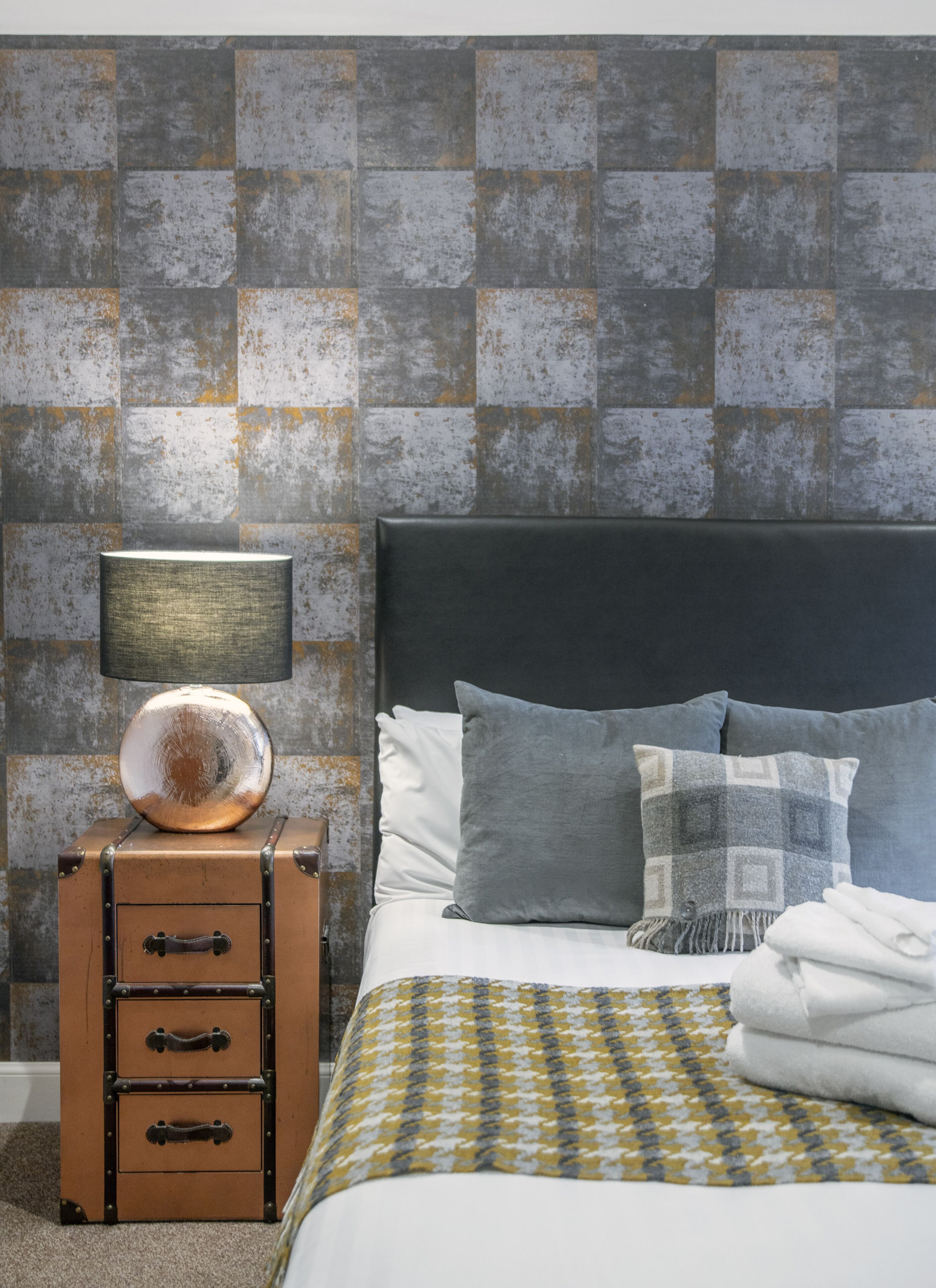 A neatly made bed with a patterned throw, a lamp on a vintage-style dresser, and a textured wall.