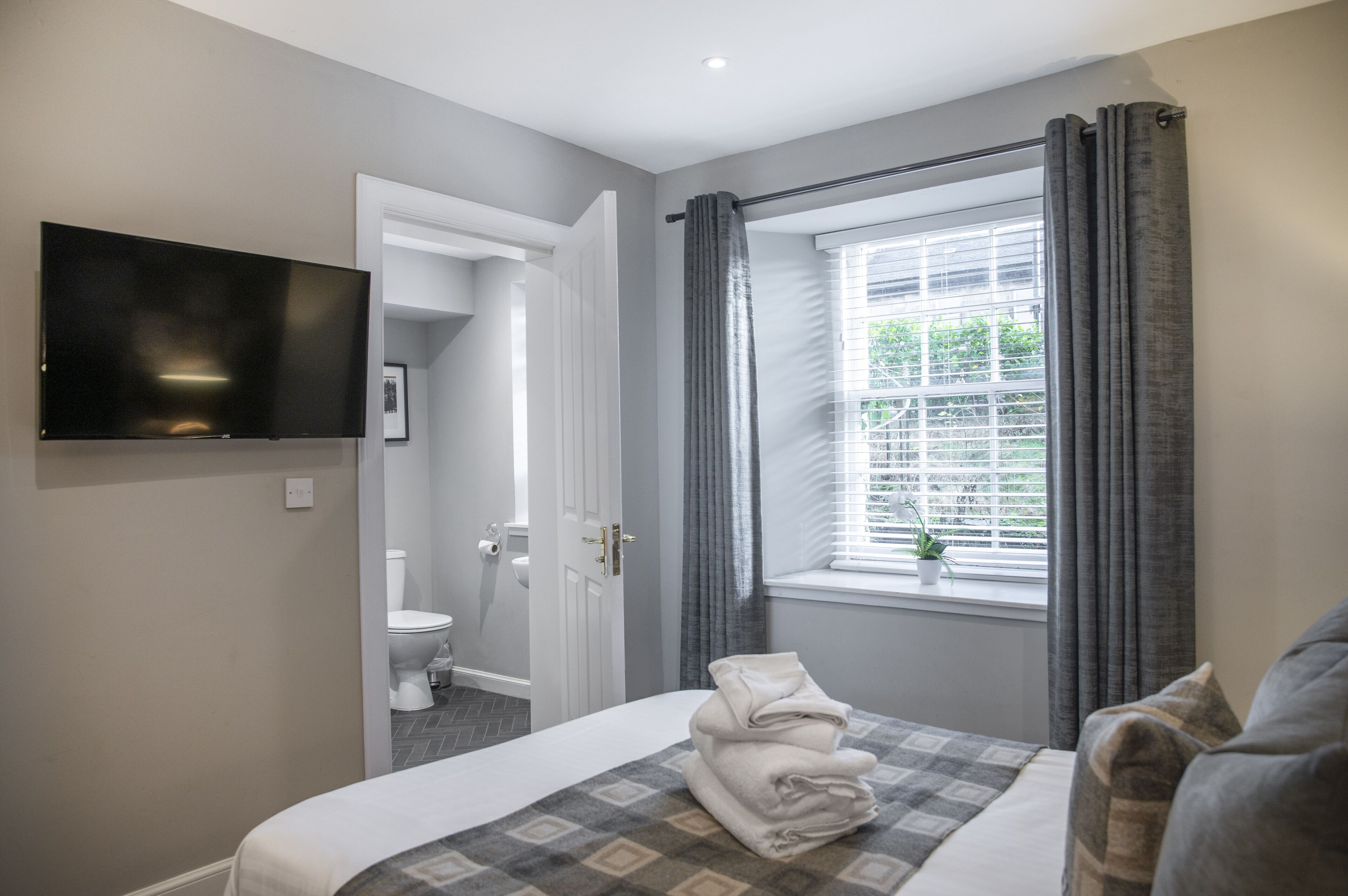 A hotel room with a bed, a TV mounted on the wall, a window with curtains, and a view of the outdoors.