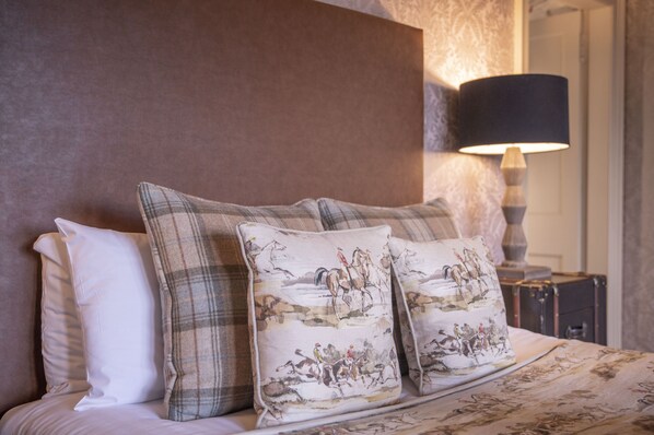 Una cama bien hecha con cabecera, dos almohadillas decorativas con escenas de equitación y una lámpara de noche.