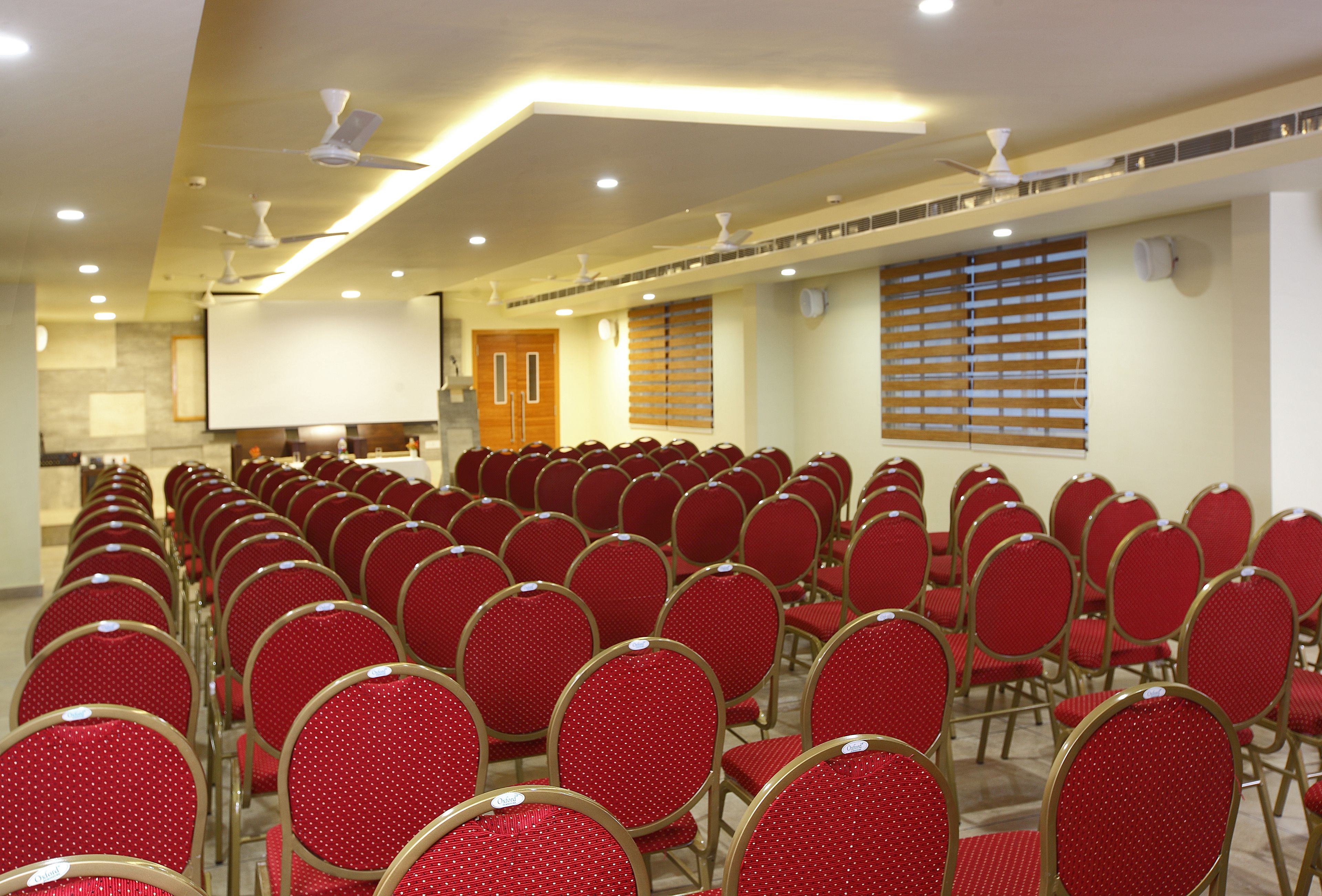 A conference room with rows of red chairs, ceiling fans, and a projector screen.