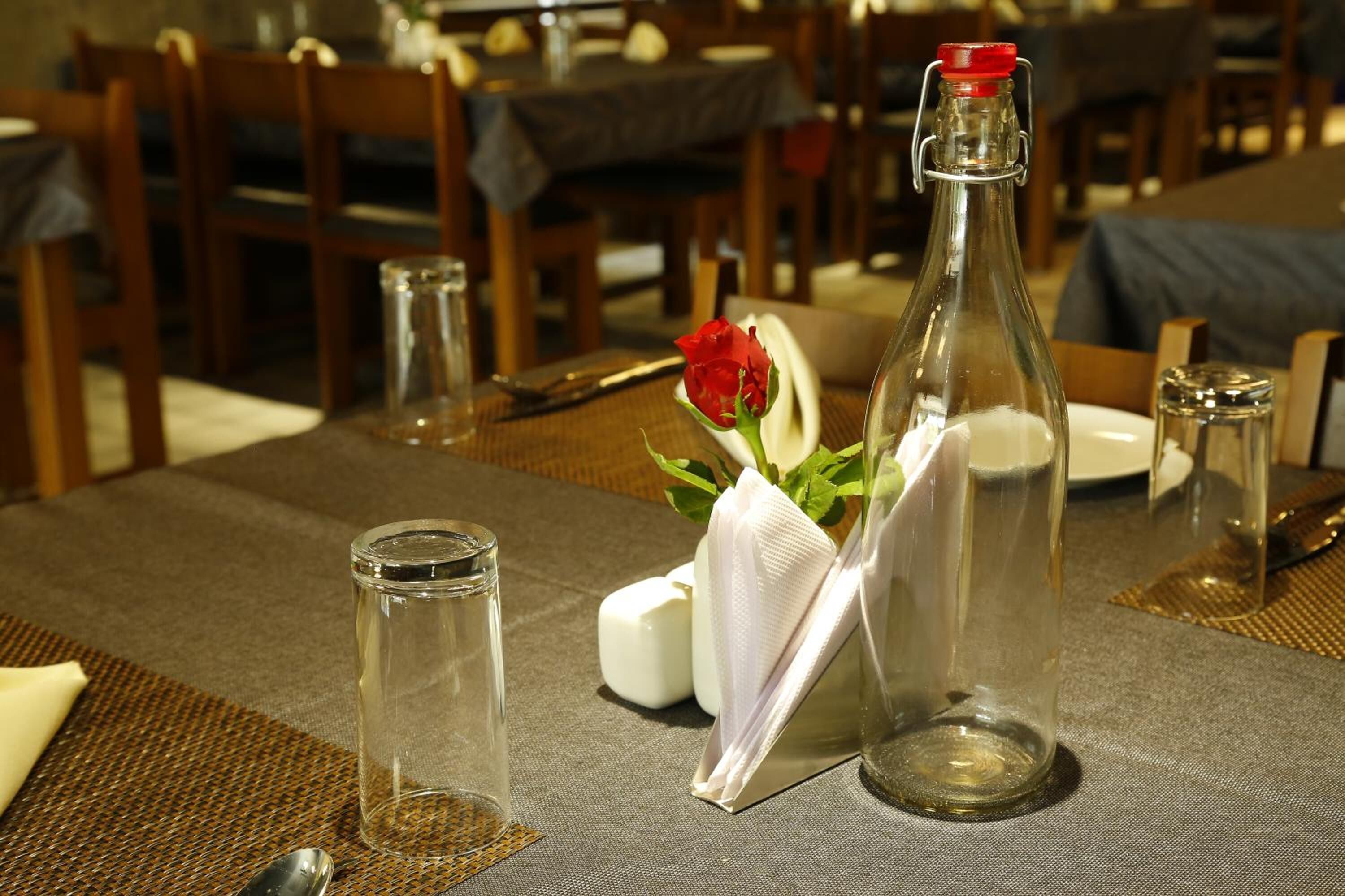 Een eettafel gedekt met een glazen fles, een witte servet, een rode roos en zout- en peperstel.