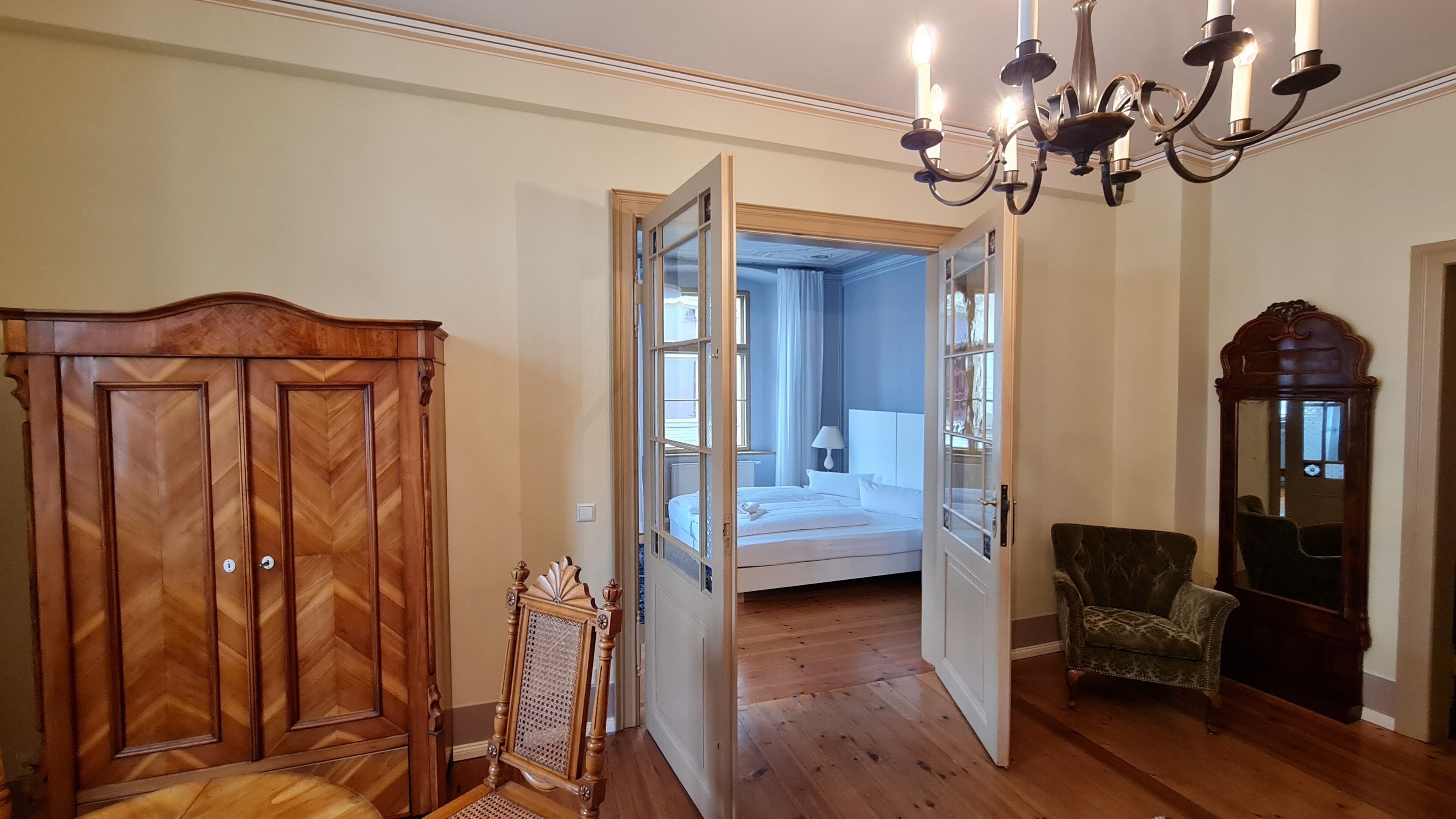 A room with a wooden wardrobe, a mirror, a chandelier, and a bed with a view to another room.