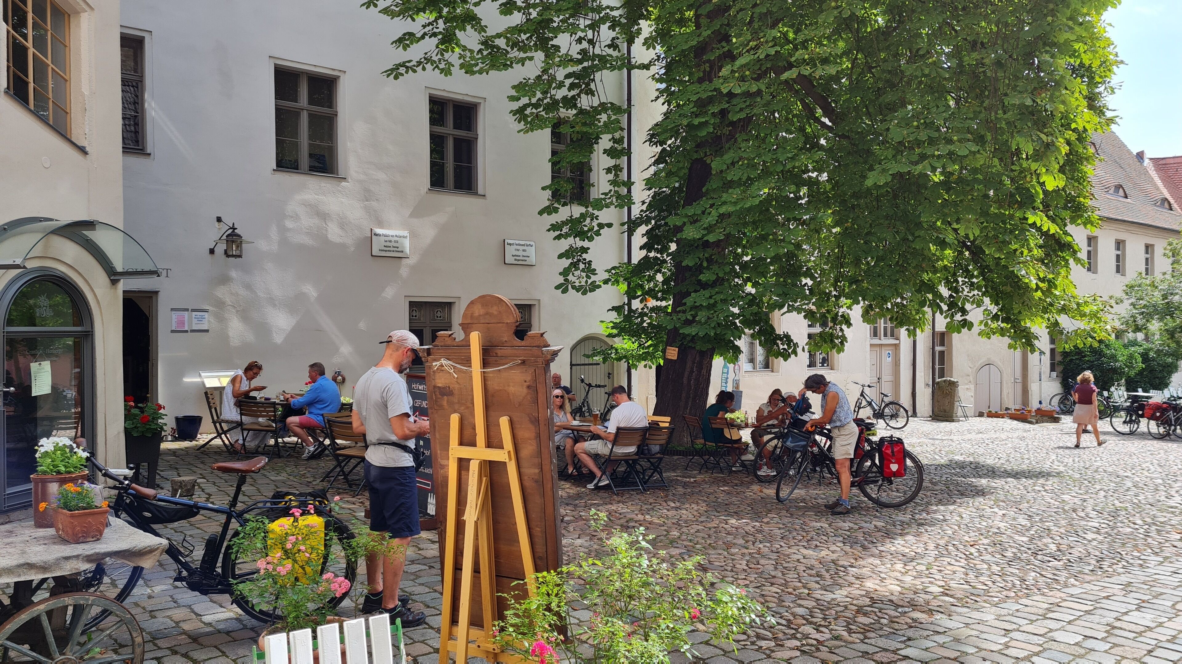 An outdoor café with tables and chairs, surrounded by cobblestone pavement and trees.