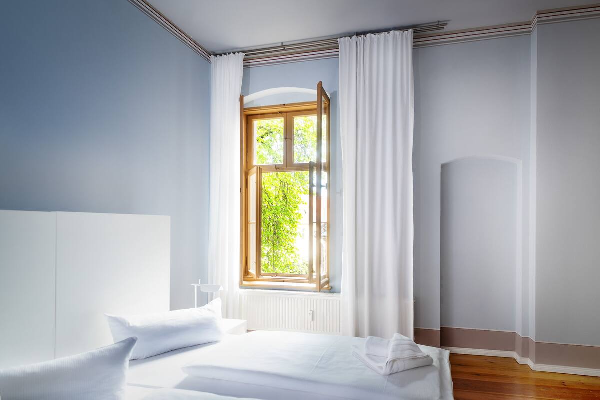 Een slaapkamer met een groot raam, witte gordijnen, een houten vloer en een wit bed.