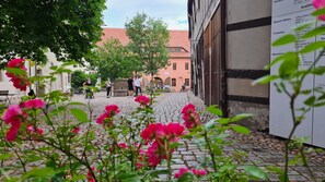 En brostensbelagt gårdsplads med lyserøde blomster i forgrunden, en bygning med rødt tag i baggrunden, og mennesker der går.