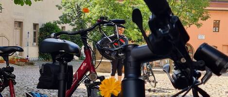 Uma bicicleta com uma flor no guidão, estacionada em uma rua de paralelepípedos com edifícios ao fundo.