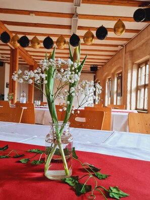 Et bord med en glassvase der indeholder hvide blomster og grønne blade, placeret på en rød dug i et rum med træbjælker og store vinduer.