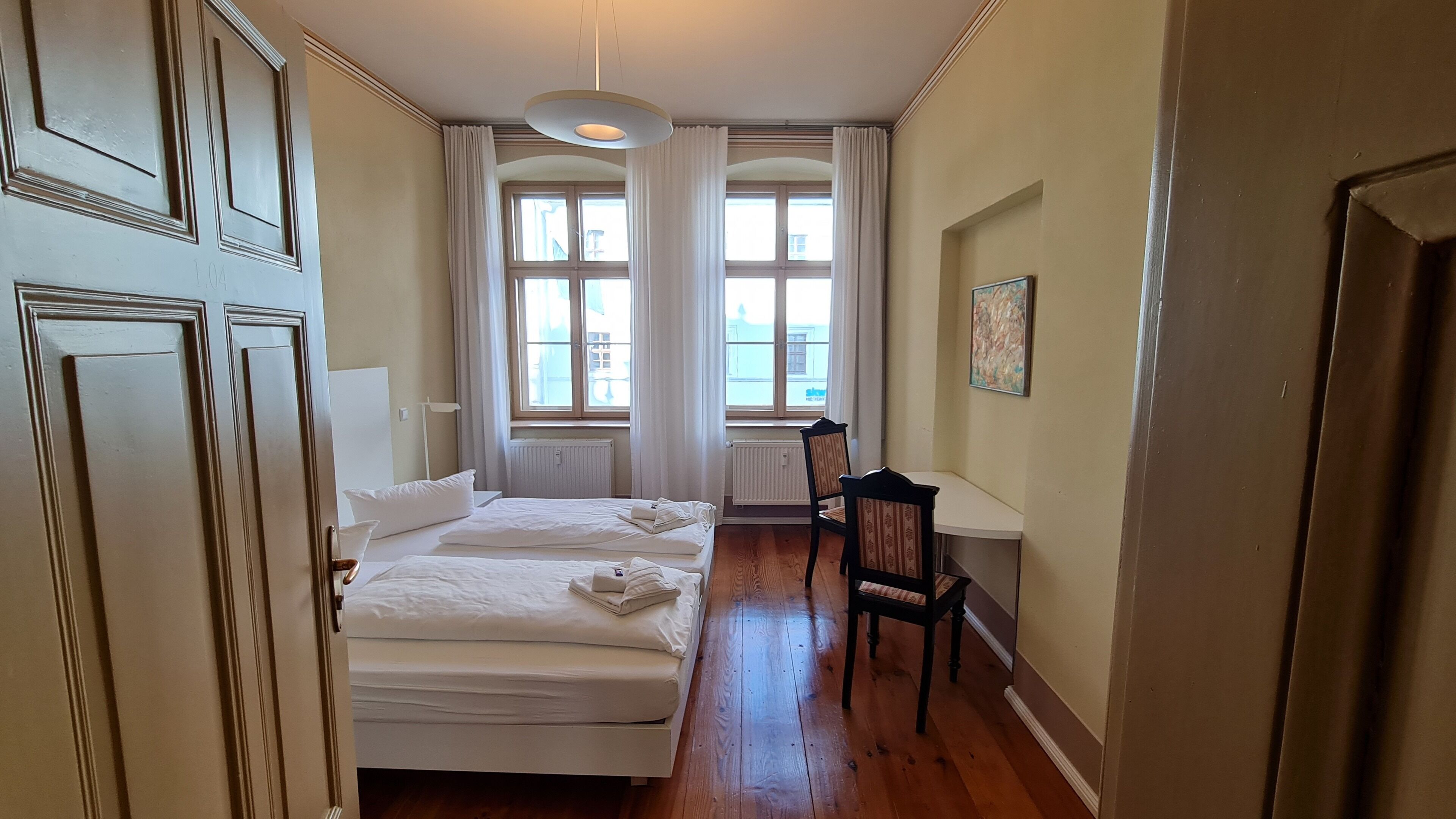 A room with two beds, a chair, a radiator, and large windows.
