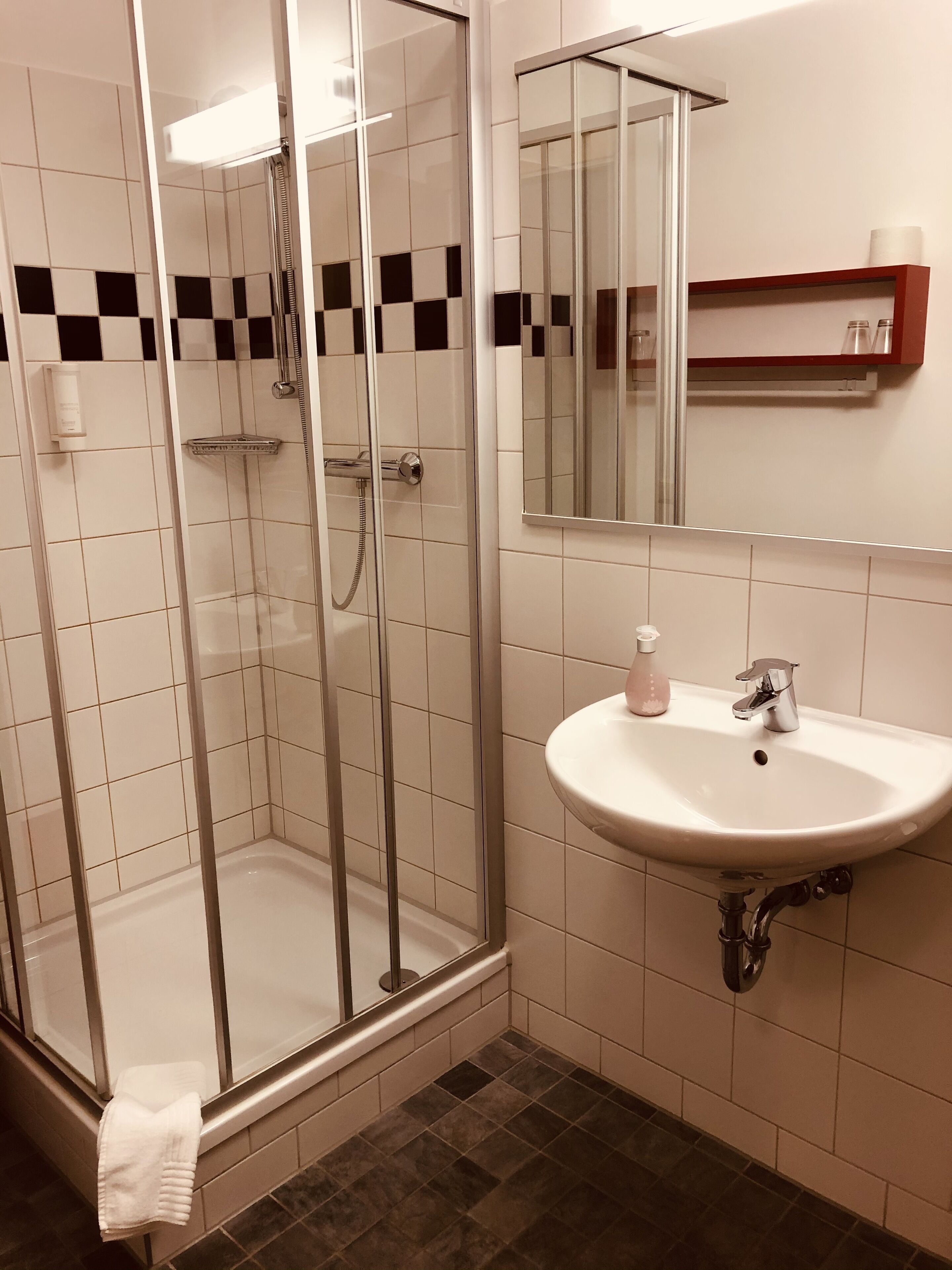 Un baño con ducha de vidrio, un lavamanos blanco con un espejo arriba y un pequeño estante con artículos de aseo.