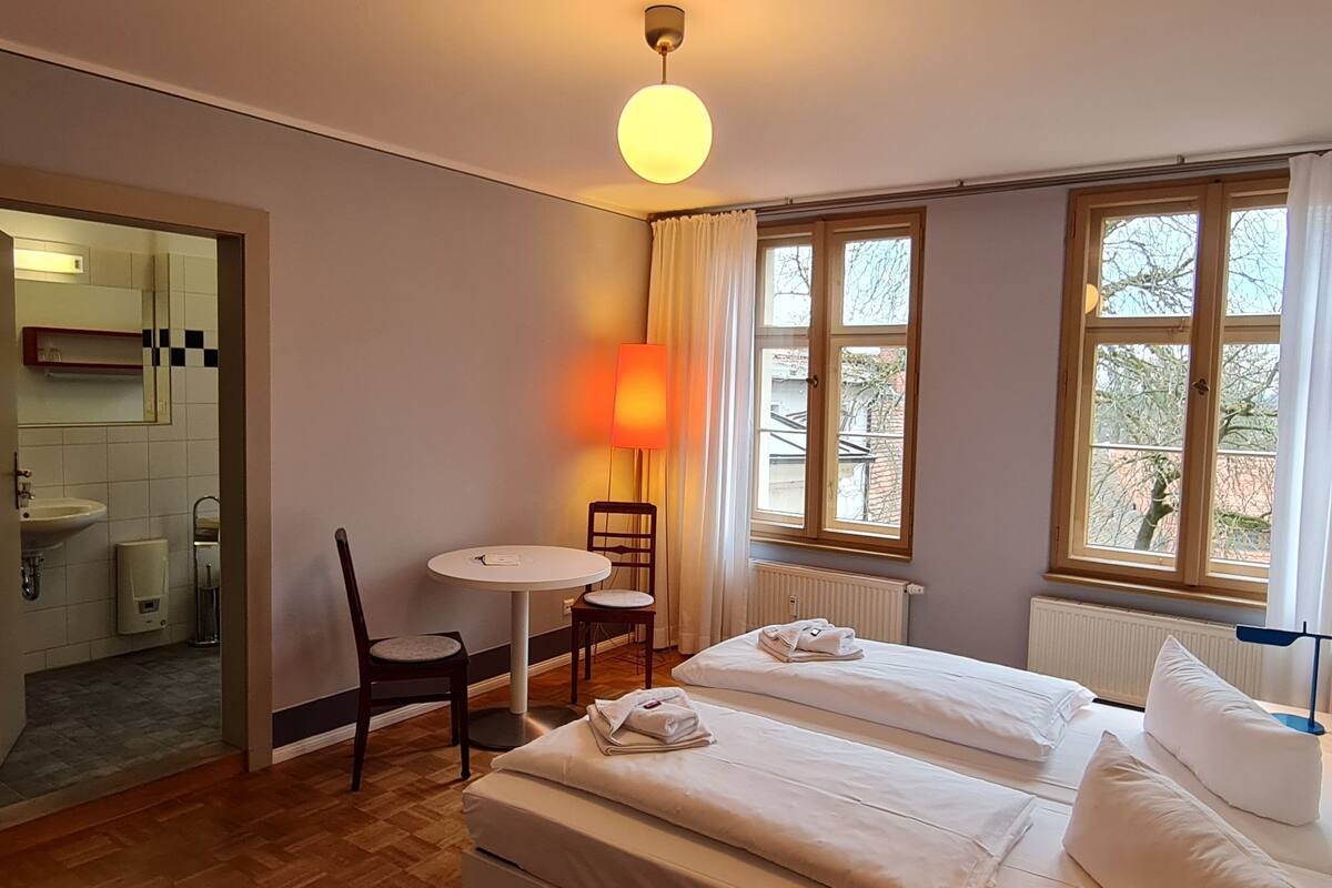 Een hotelkamer met twee bedden, een tafeltje, een stoel, een lamp en uitzicht op bomen door het raam.