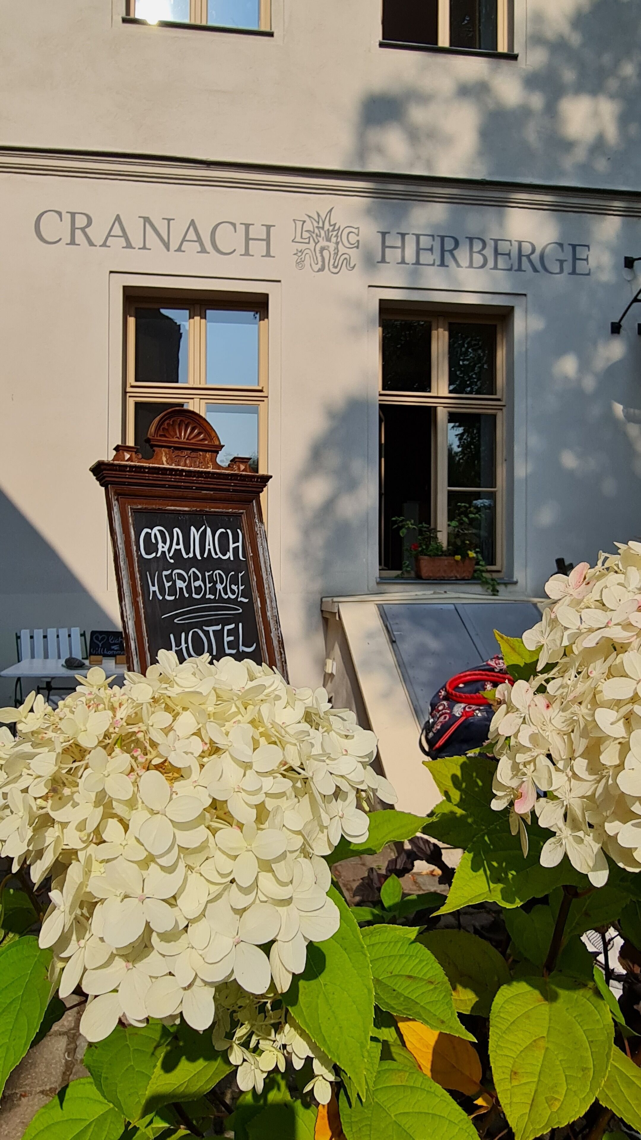 A hotel sign with "CRANACH HERBERGE HOTEL" and a signboard for "CRANACH HERBERGE HOTEL" in front of a building with windows and a staircase.
