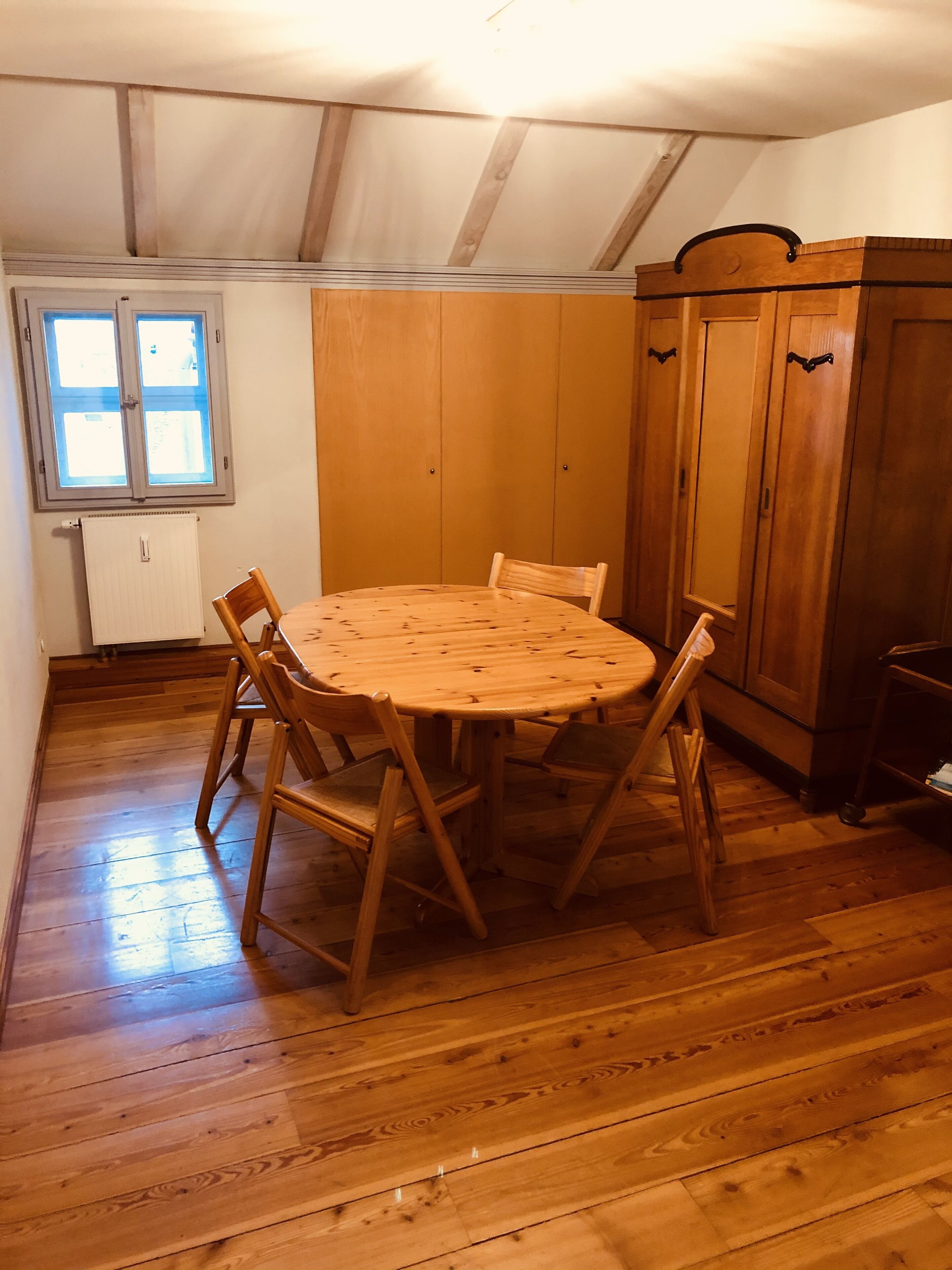 Een kamer met een houten eettafel en stoelen, een houten kledingkast, en een raam met jaloezieën.