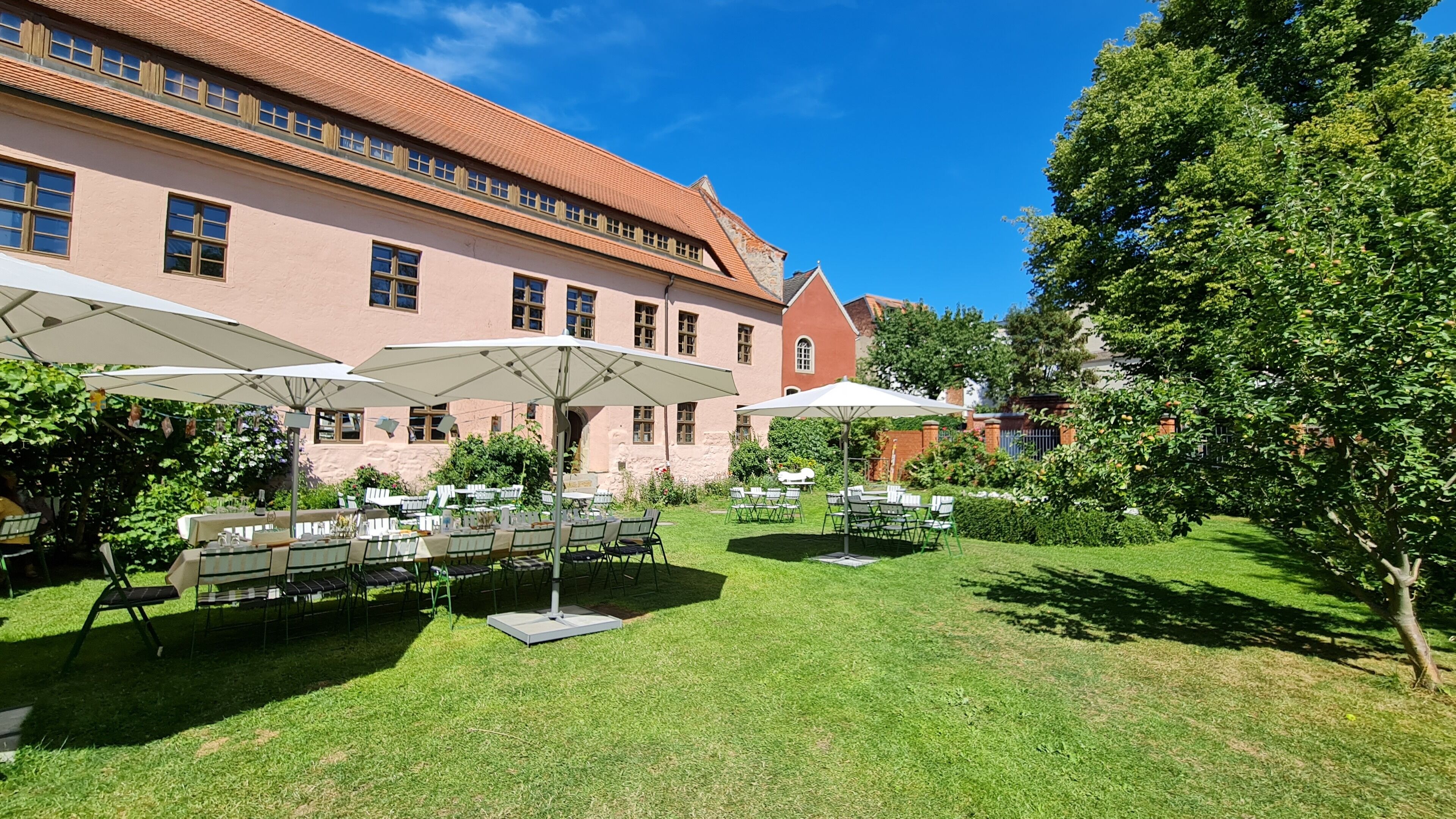 An outdoor patio with tables and umbrellas, surrounded by a well-maintained lawn and a large building with a red-tiled roof.