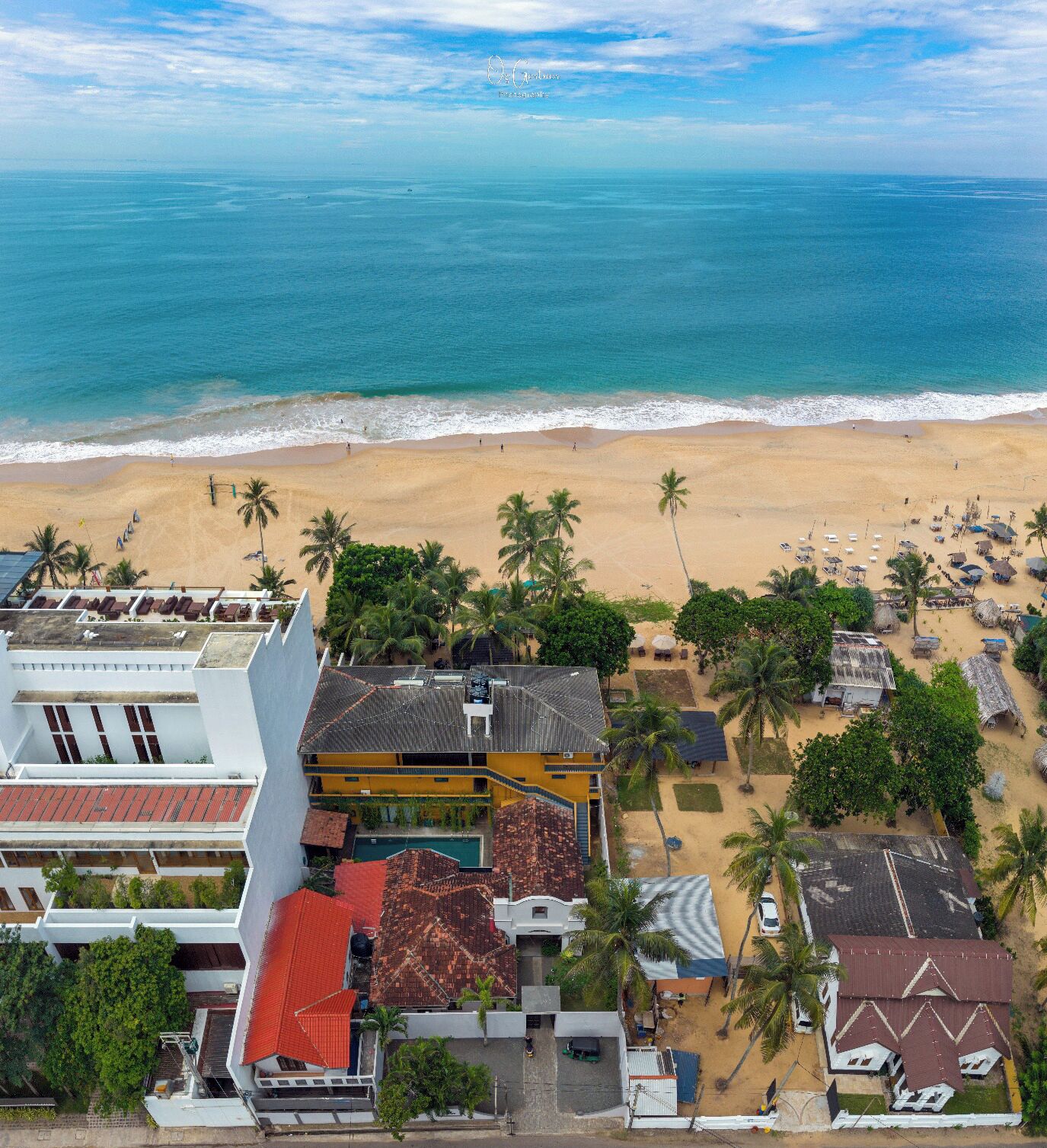 An aerial view of a beachside property with a large building, a swimming pool, and a beach with palm trees and umbrellas.