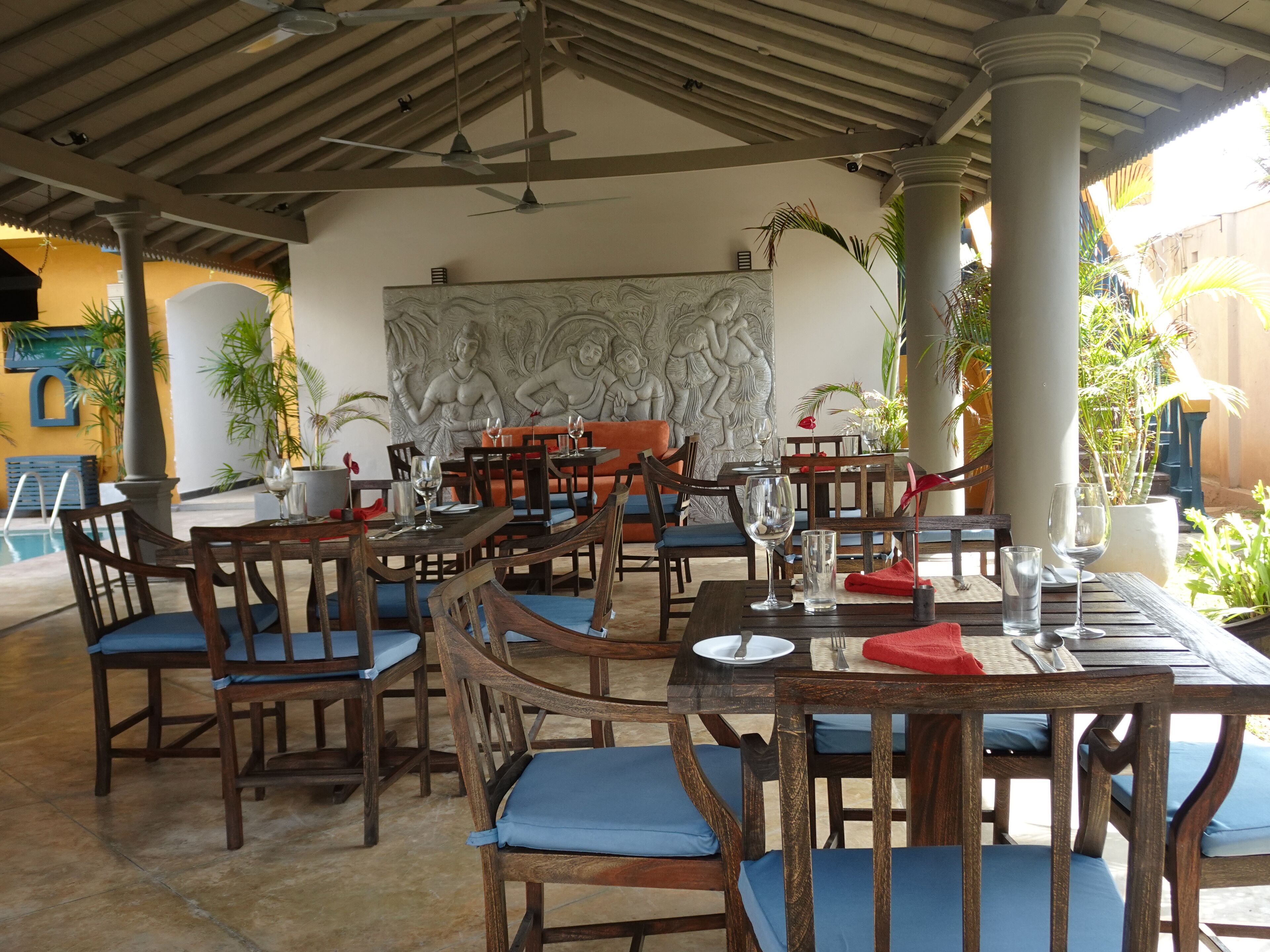 Un espacio de comedor al aire libre con mesas y sillas de madera, una pared decorativa con esculturas en relieve y una piscina al fondo.