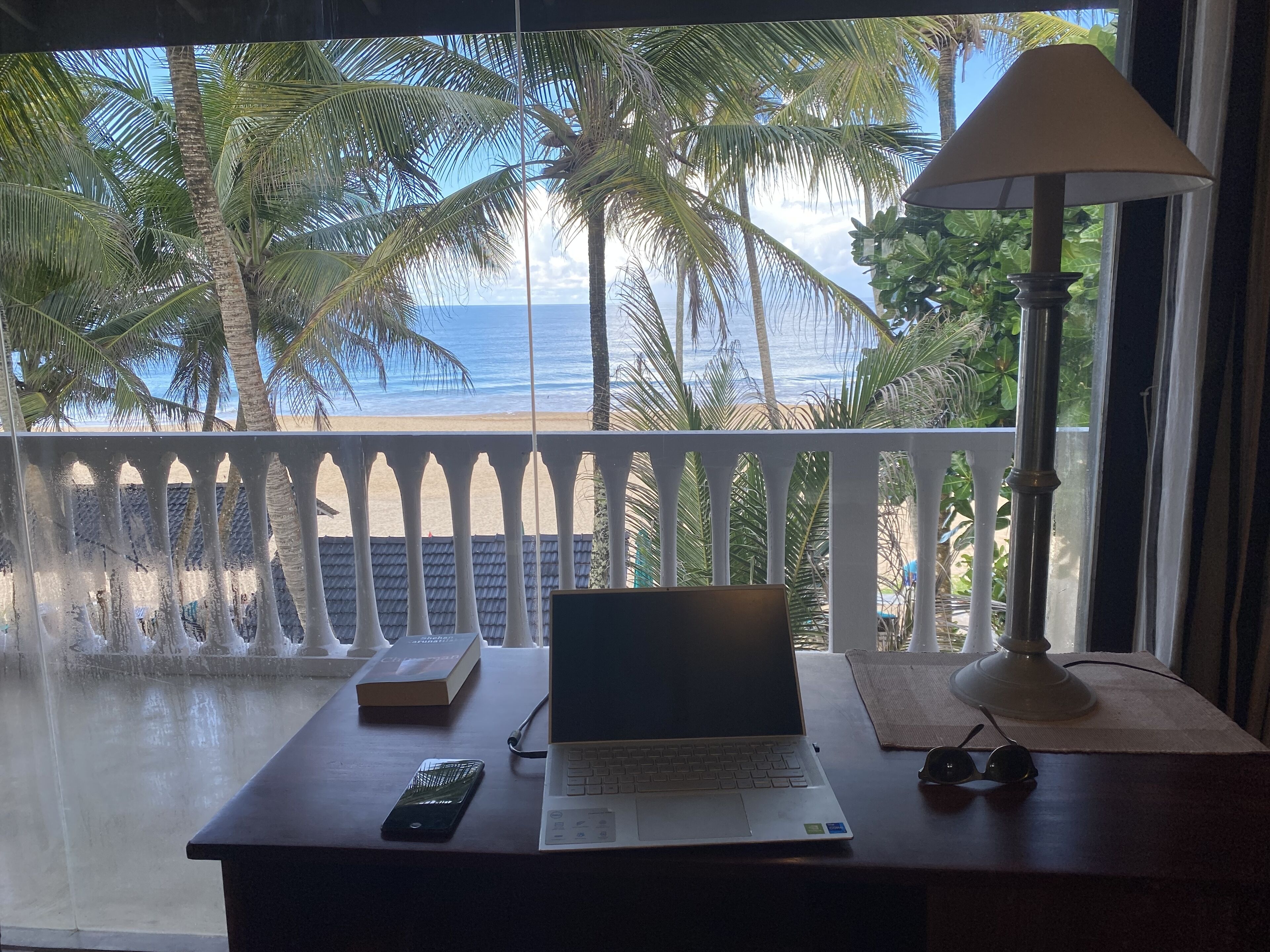 Un escritorio con una computadora portátil, un libro y un teléfono inteligente, con vista a una playa con palmeras y el océano.