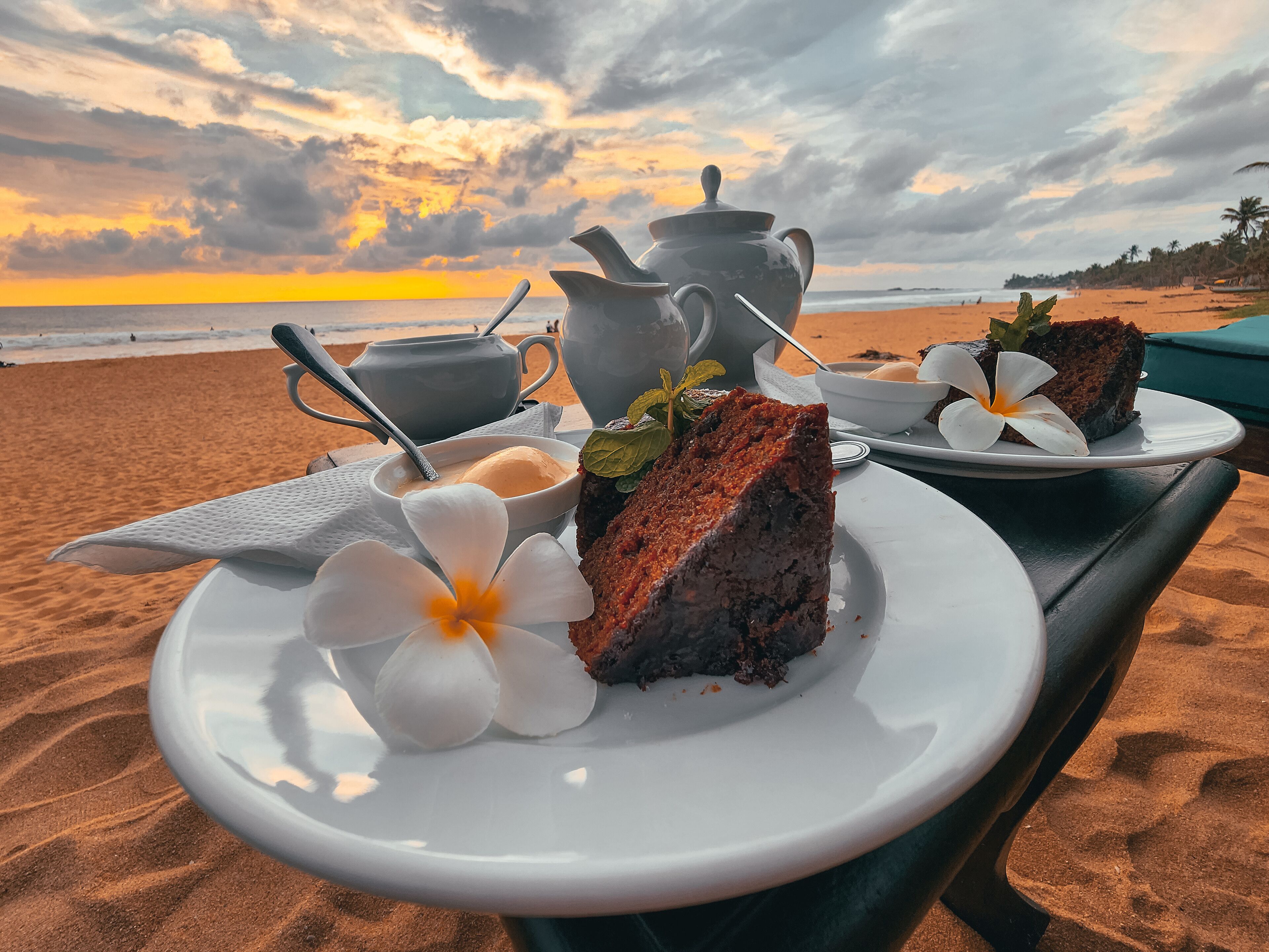 Una mesa en la playa con un pastel, una tetera y una taza de café, con el atardecer de fondo.