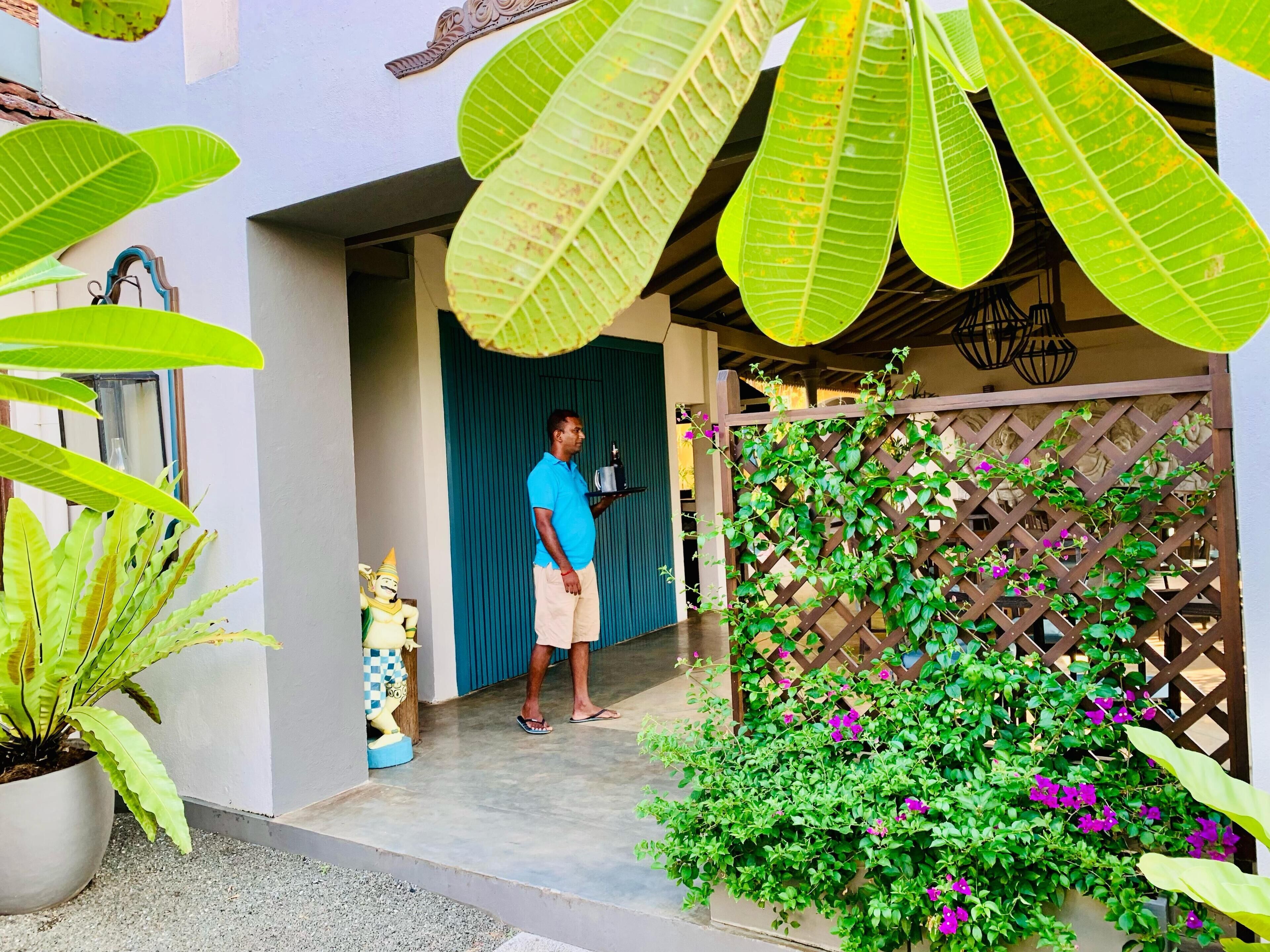 A man standing at a doorway holding a tray, with a decorative statue and potted plants in the foreground.