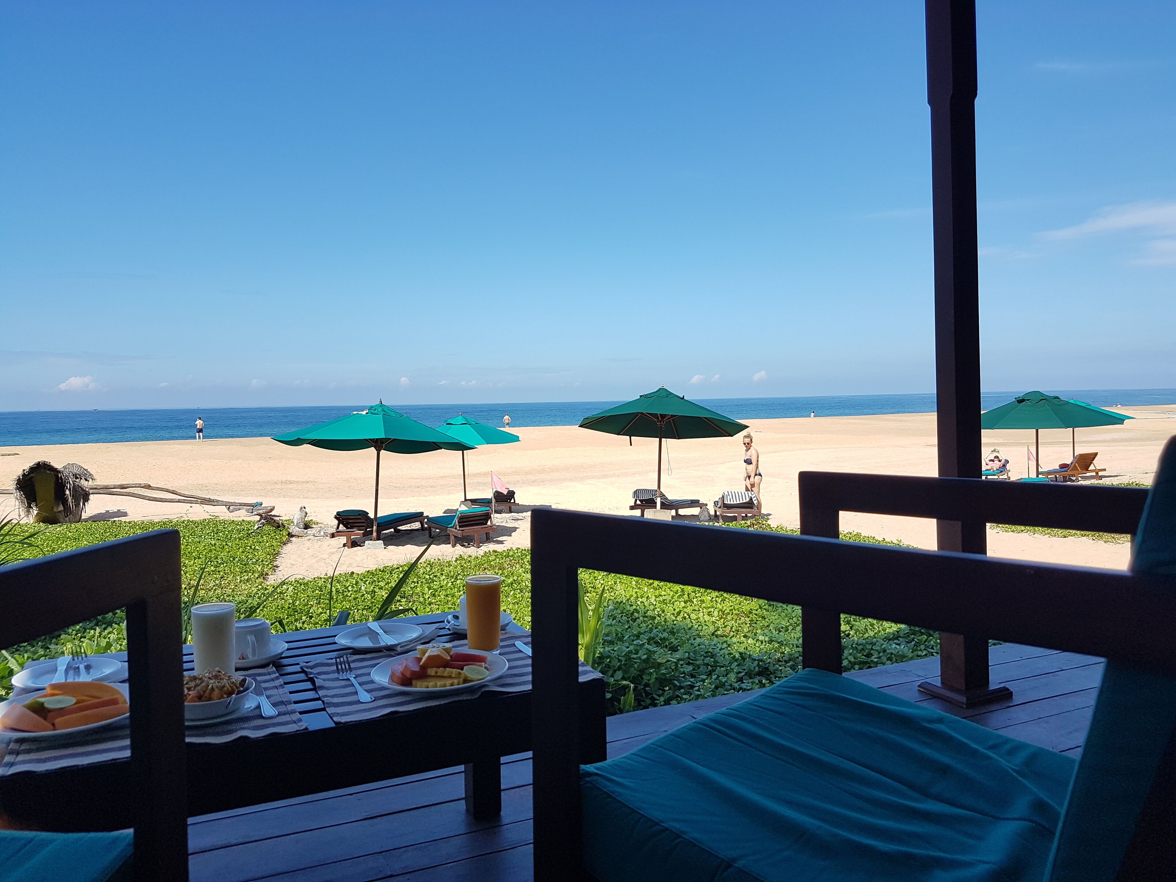 A beach view from a covered deck with dining tables, chairs, and umbrellas.