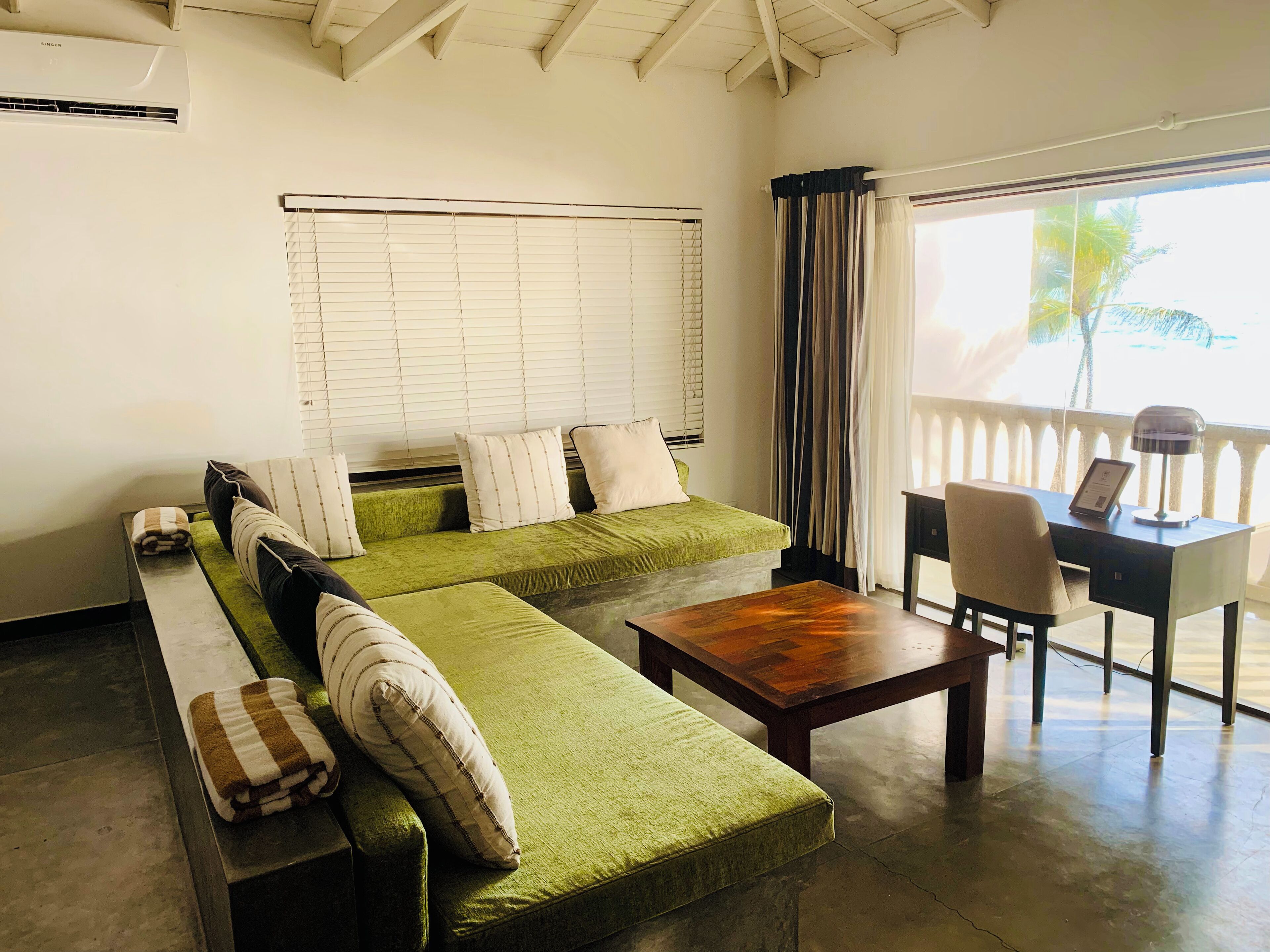 A room with a green sofa, a wooden table, a desk with a lamp, and a window with curtains.