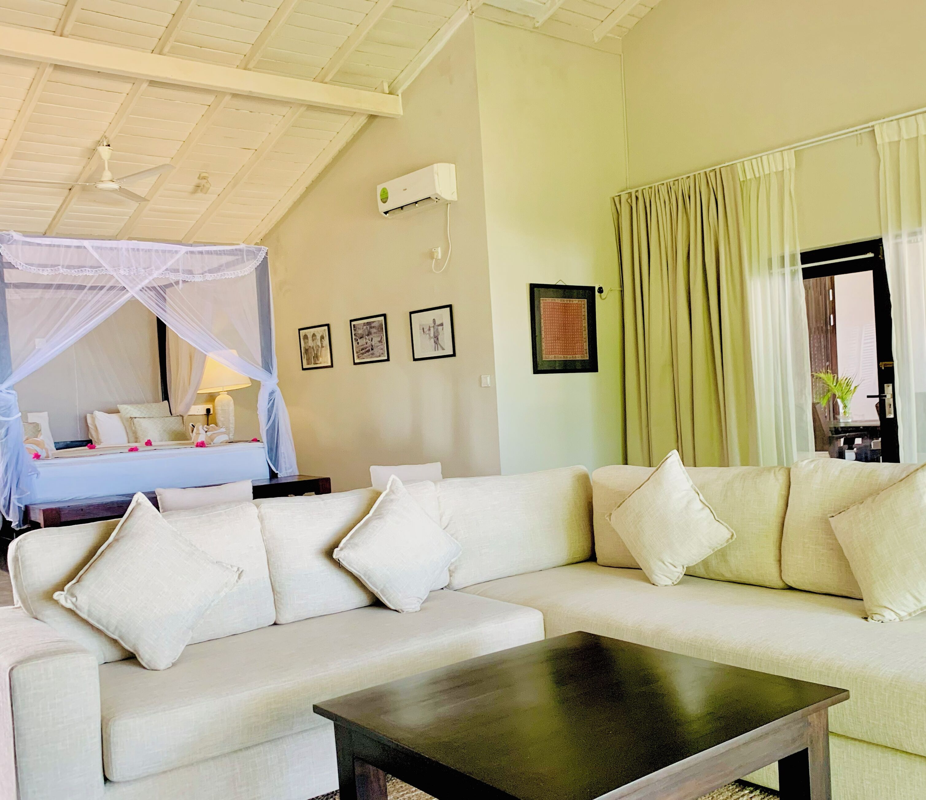 Une pièce avec un canapé blanc, une table basse noire, un ventilateur de plafond, un lit à baldaquin et un climatiseur fixé au mur.