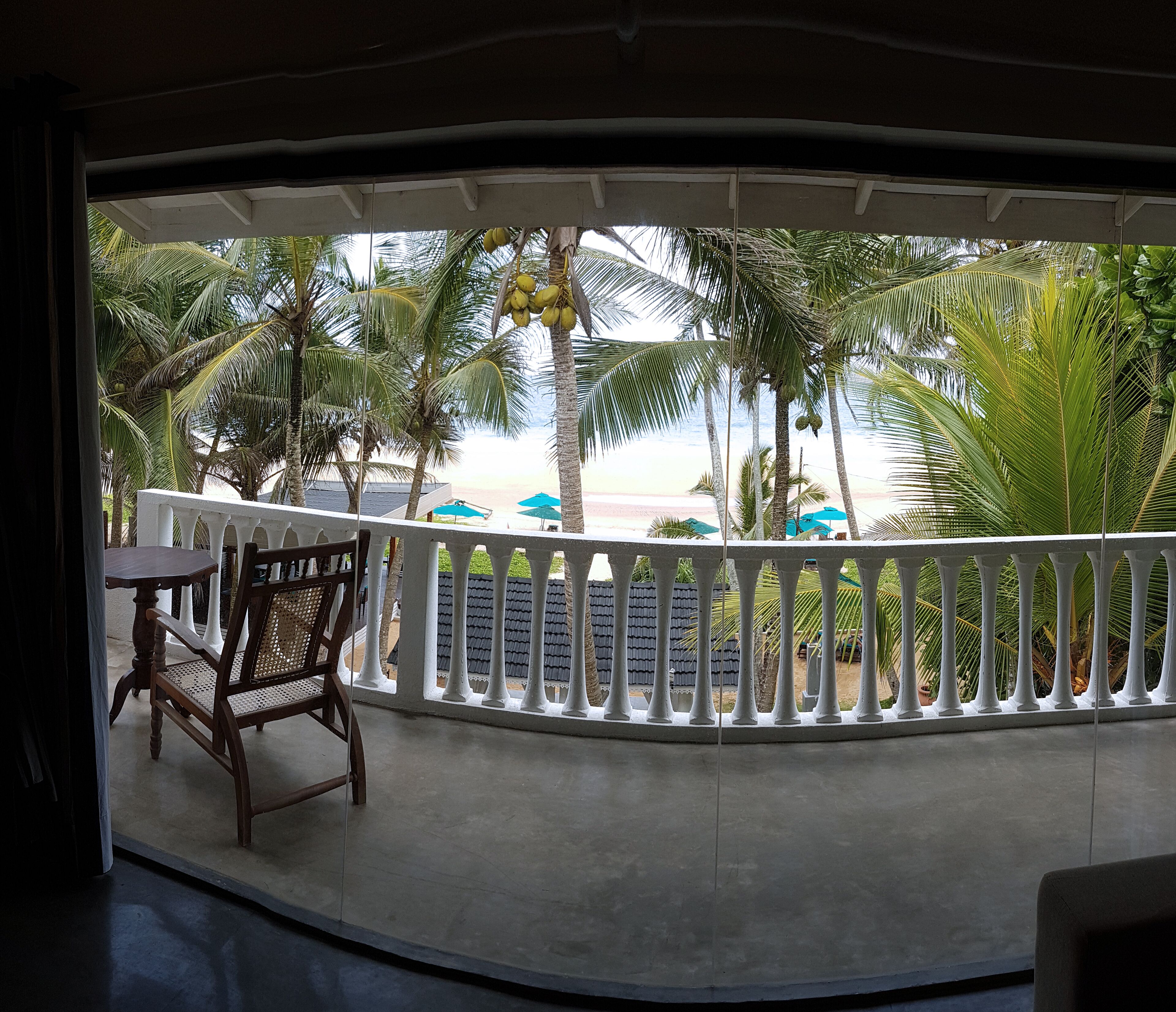 Een balkon met een witte reling, een houten stoel en een tafel. Palmbomen en een strand zijn in de achtergrond zichtbaar.