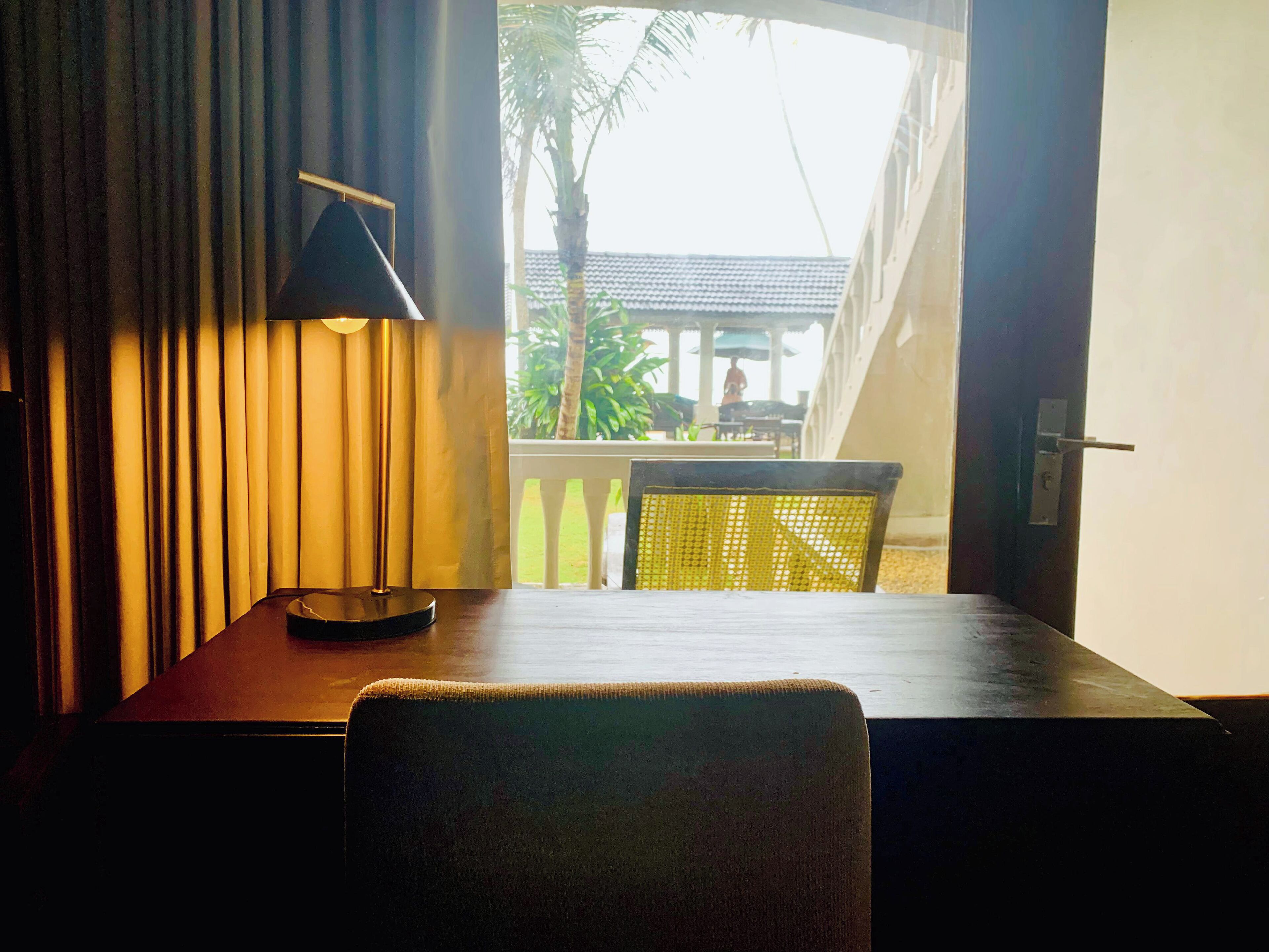 Ein Zimmer mit Tisch, Stuhl, einer Lampe und Blick auf einen Balkon mit einer Palme.