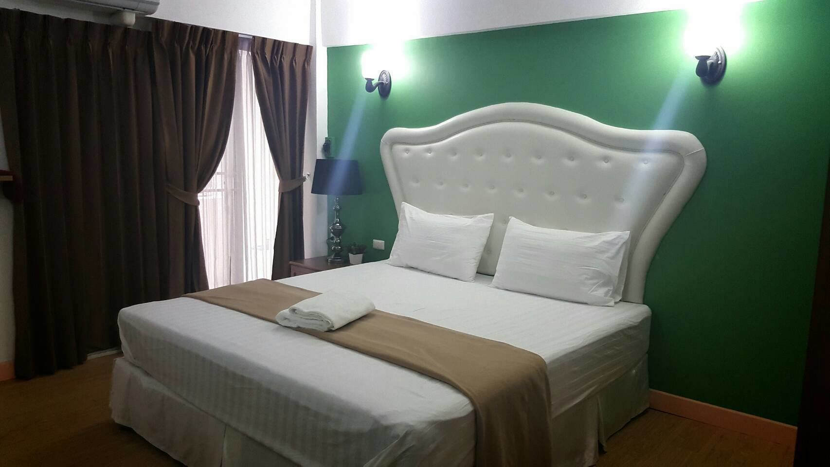 A neatly made bed with white linens and a brown blanket, a green wall, and two wall-mounted light fixtures.