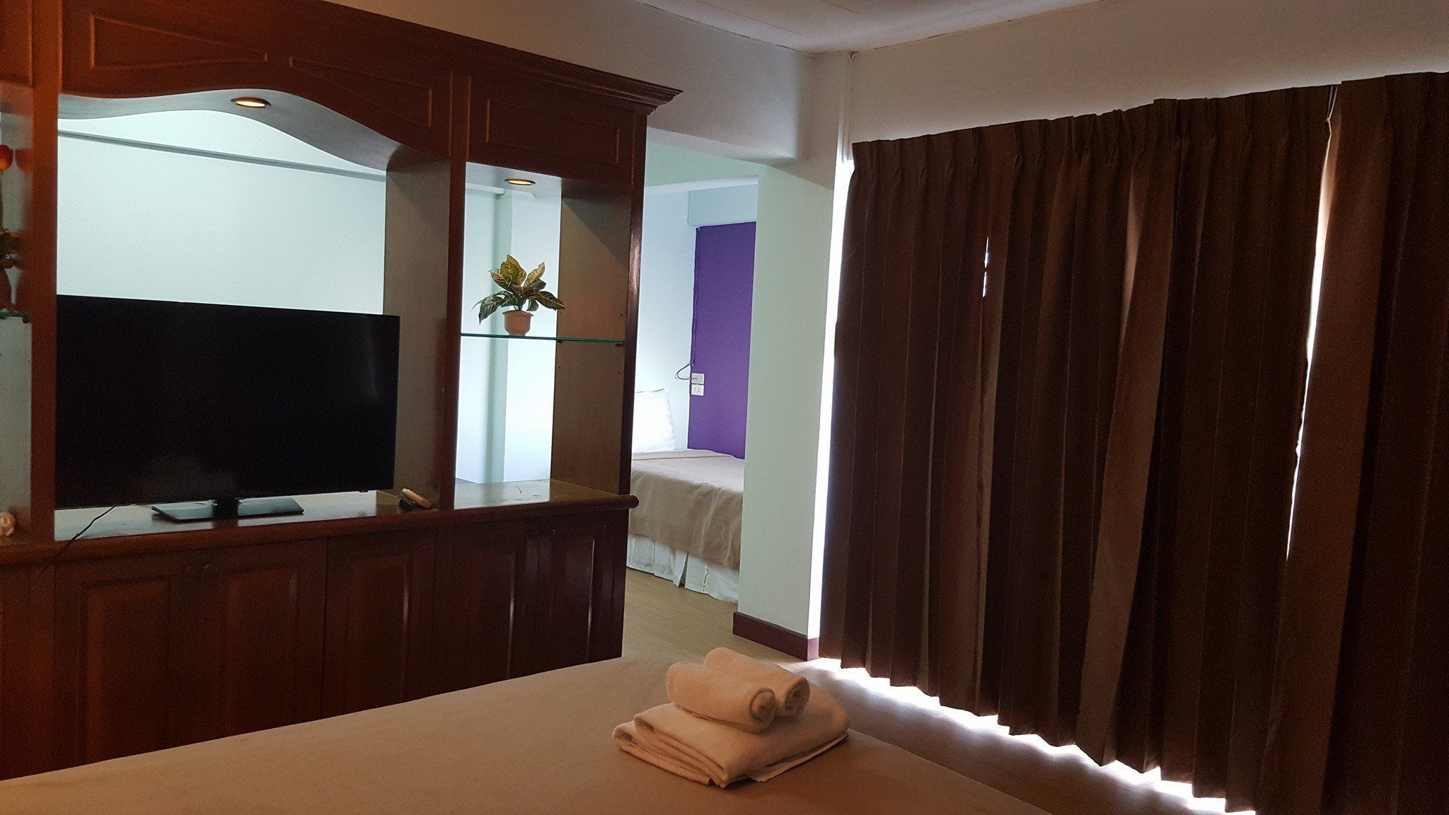 Una camera d'albergo con una televisione, un tavolino in vetro con una pianta in vaso, un letto con copriletto color beige e tende marroni.