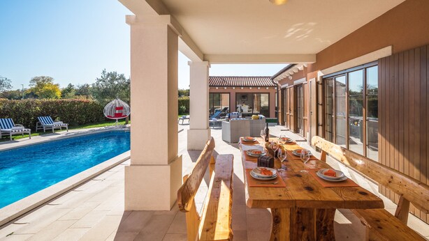 A poolside dining area with a wooden table set for a meal, benches, and a view of the pool and house in the background. - Villa Kanco (Barban)