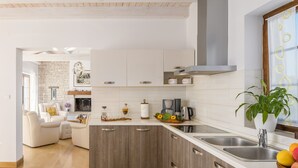 A modern kitchen with wooden cabinets, a white countertop, and a built-in stove. There is a dining area with a white sofa and a coffee machine.