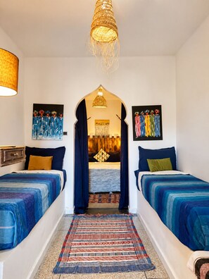 A hotel room with two beds, a patterned rug, a hanging light fixture, and two paintings on the walls. - Yousurf Surf House (Essaouira)