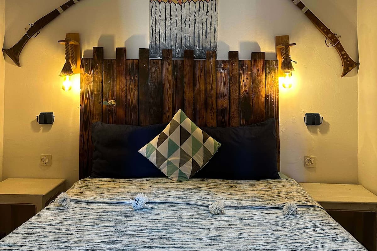 A bedroom with a bed, a wooden headboard, two wall-mounted lights, and two decorative axes.