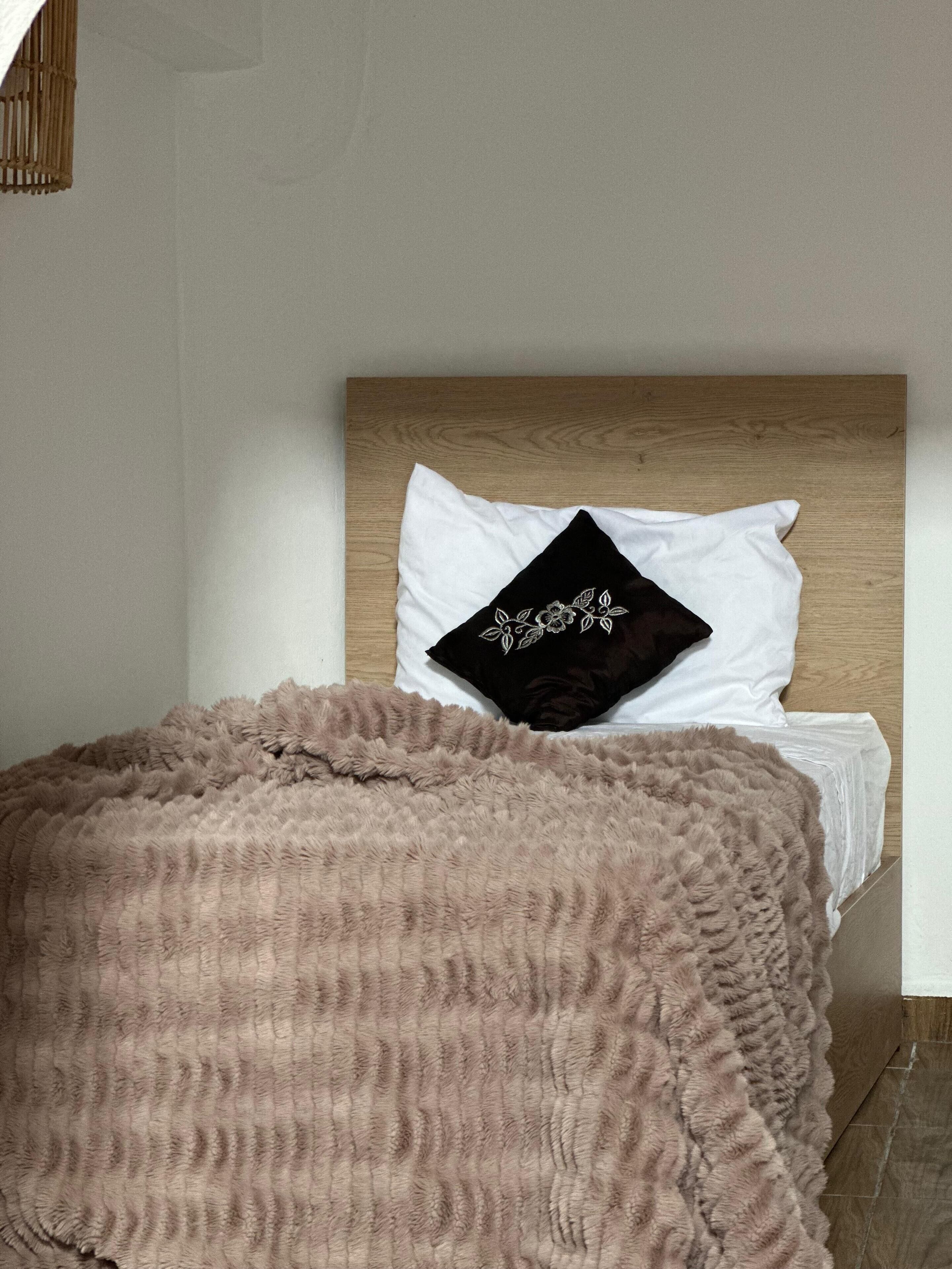Una cama con cabecera de madera, almohadas blancas y una almohada negra con bordado floral. Una manta marrón suave cubre la cama.