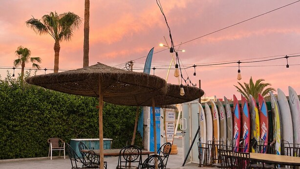 A beachside restaurant with outdoor seating, palm trees, and surfboards. - Yousurf Surf House (Essaouira)
