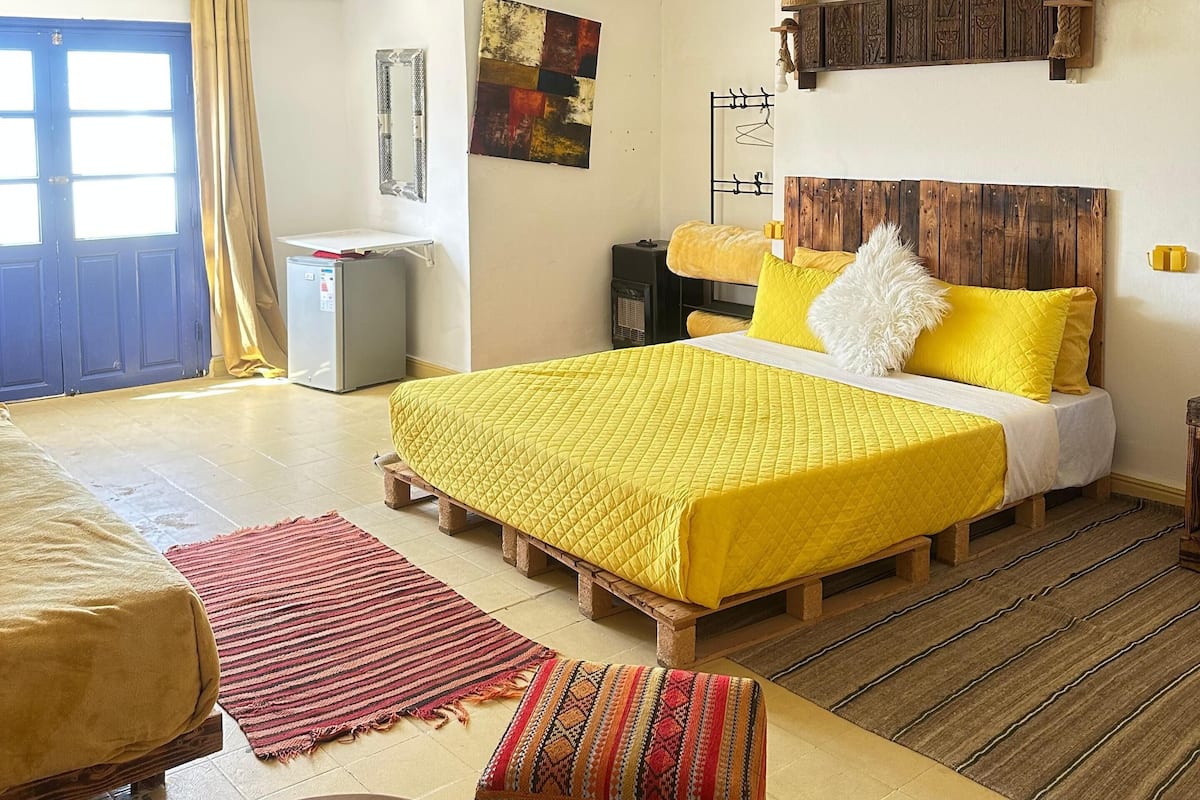 A bedroom with a bed, a small table, a bench, a heater, and a door.