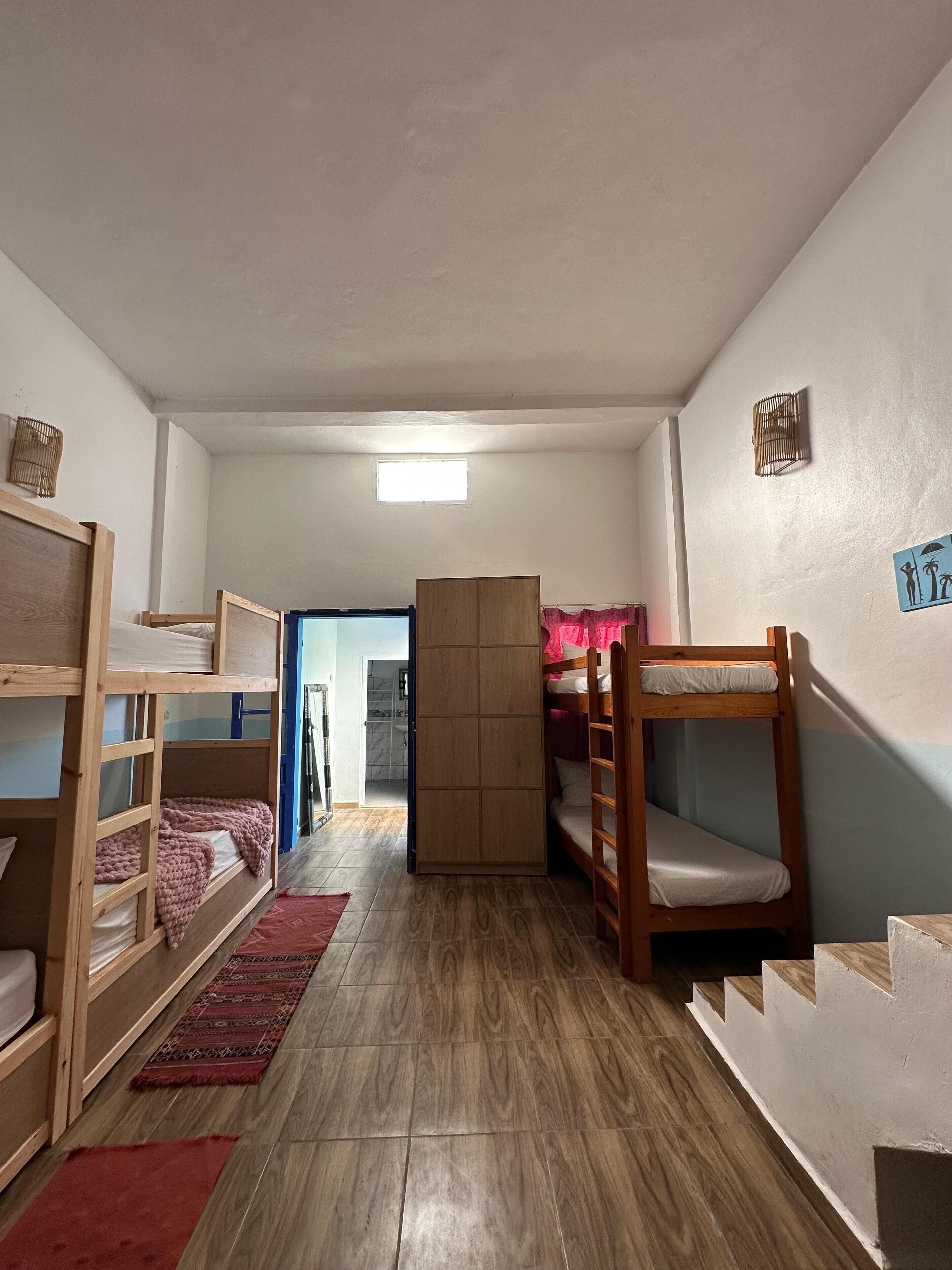 Een studentenkamer met stapelbedden, een garderobe, een klein raam en een deur naar een andere kamer.