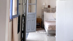 A narrow hallway with a door leading to a bedroom. The room has a bed, a nightstand, and a lamp. The hallway has a white wall and a window with curtains. - Yousurf Surf House (Essaouira)