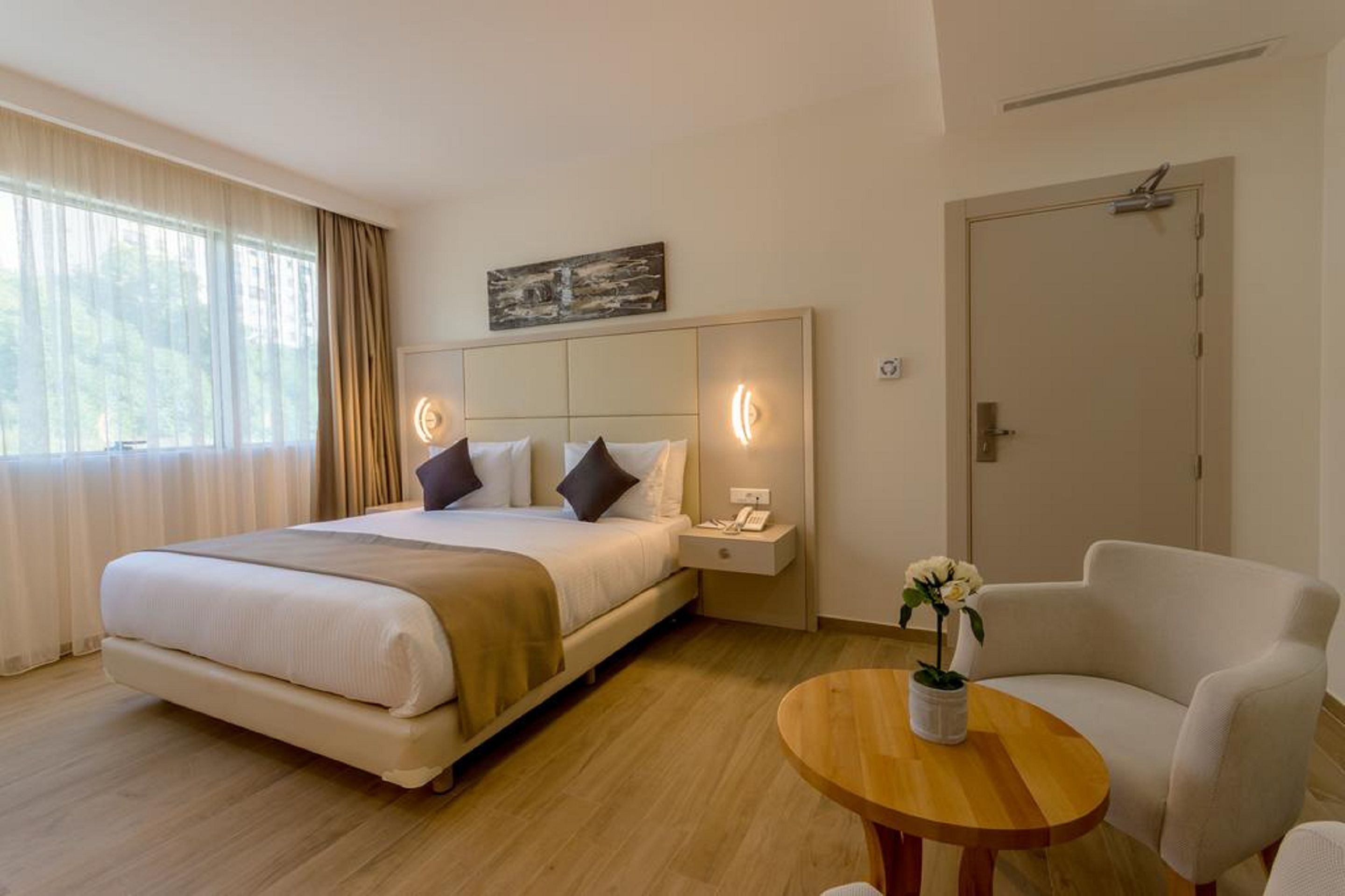 Una habitación de hotel con cama, mesita de noche, lámpara, silla y una pequeña mesa con un florero.