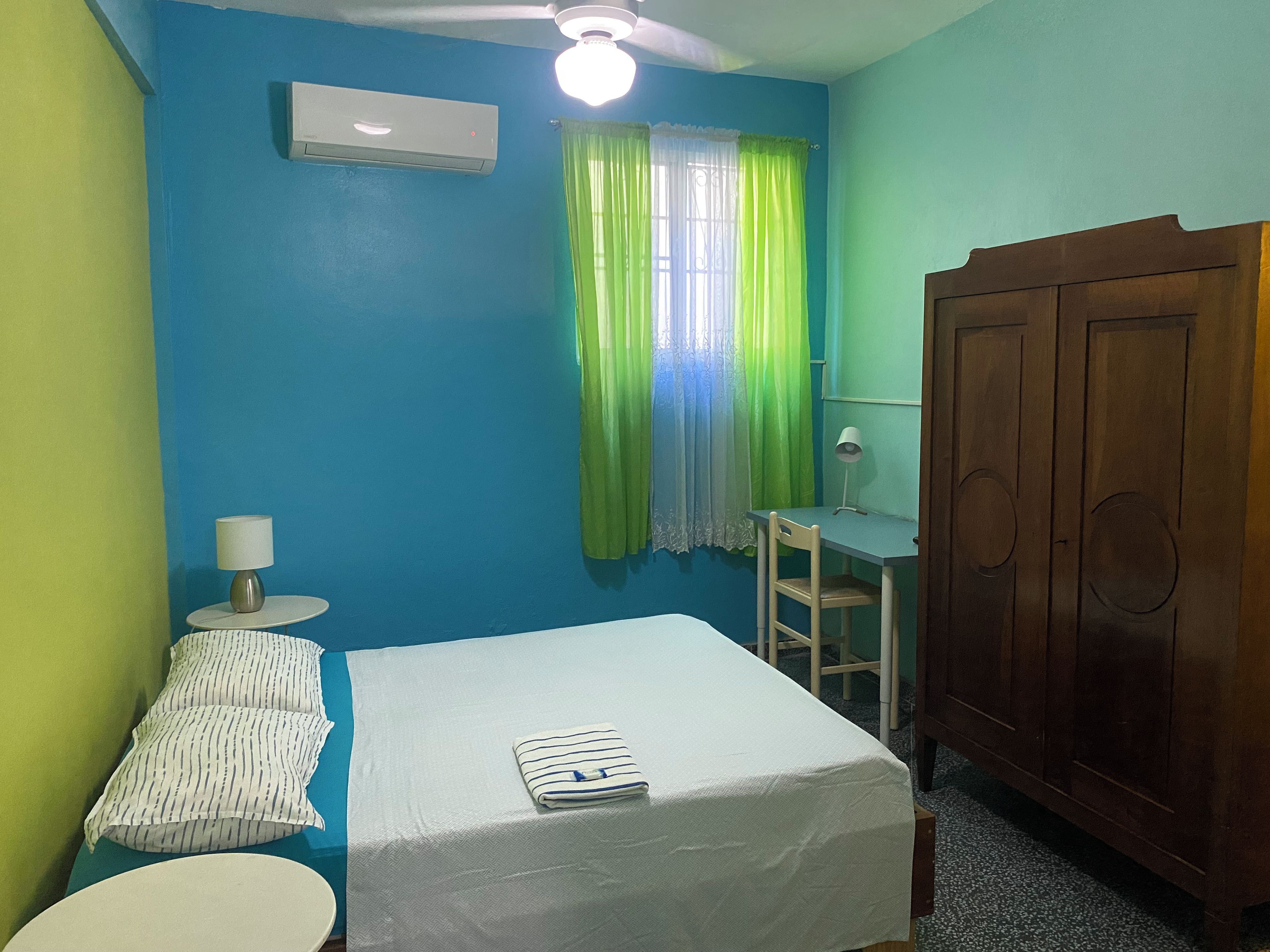 Ein Zimmer mit einem Bett, einem Schreibtisch, einem Kleiderschrank, einem Vorhangfenster und einer Klimaanlage.