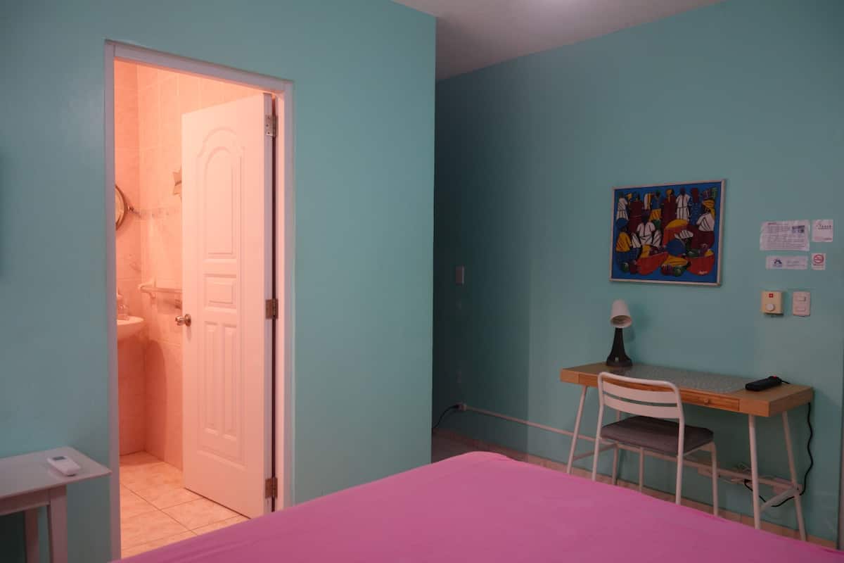 A room with a bed, a desk, and a chair. There is a door leading to a bathroom.